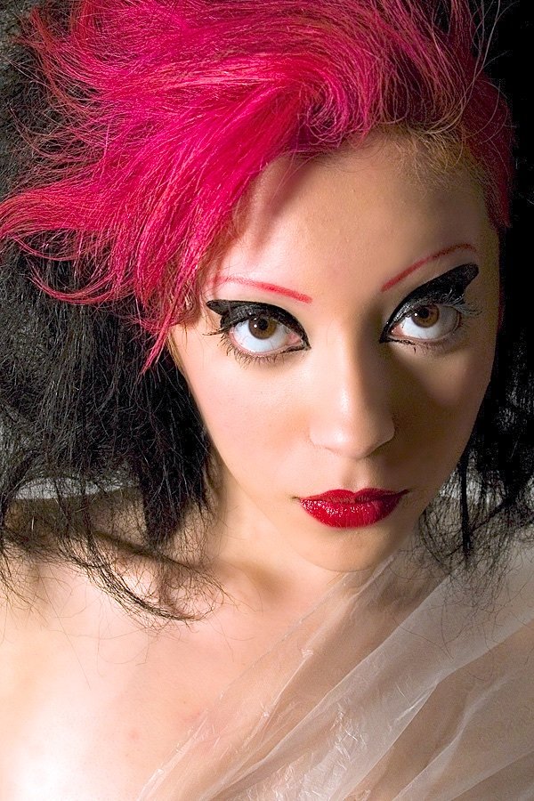 A woman with pink and black hair, bold eye makeup, and red lipstick looking up at the camera.