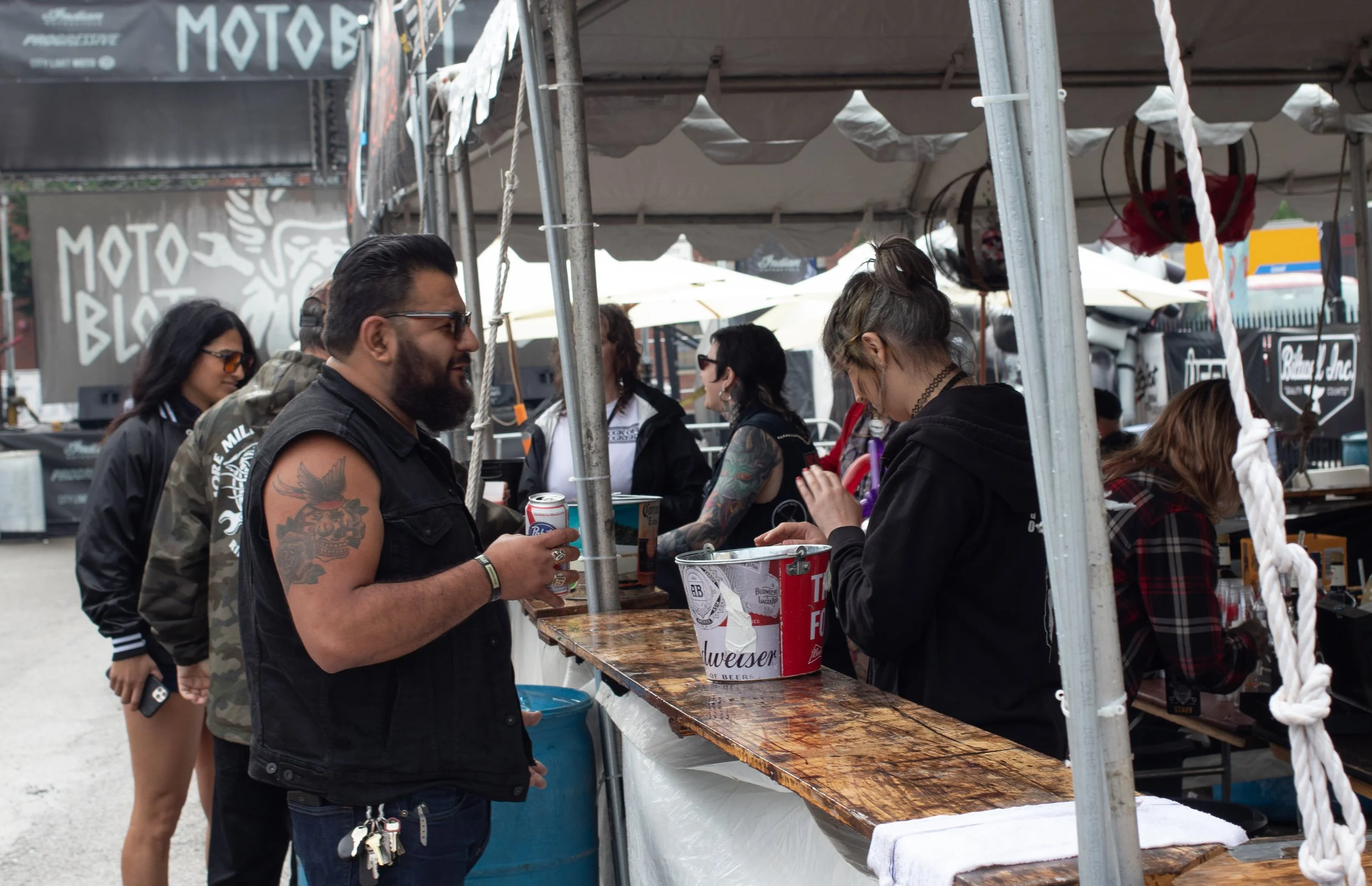 Group of people at an outdoor festival or market, some smiling and holding drinks, with a sale booth and tents in the background.