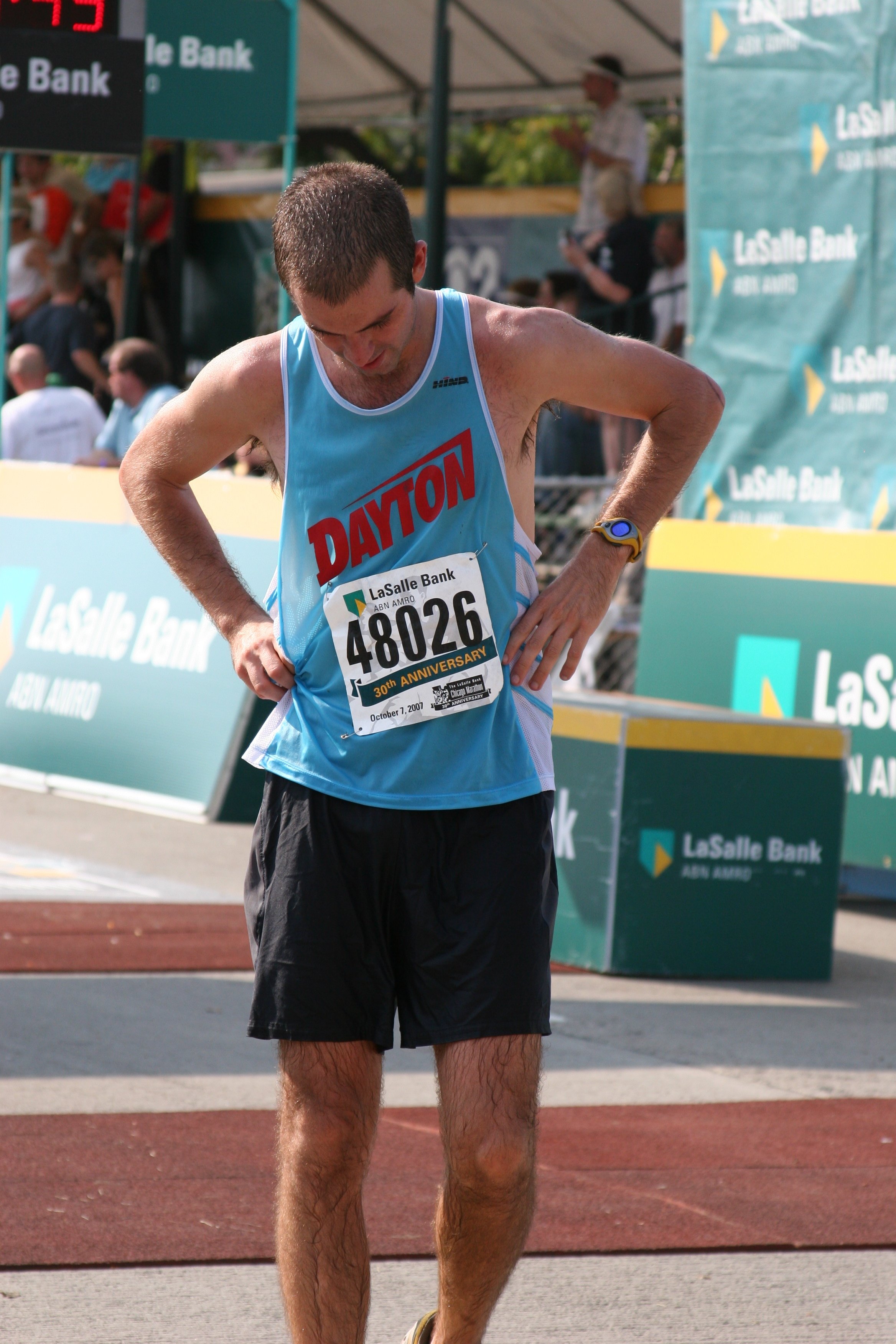 A male athlete in a blue tank top and black shorts, with bib number 48026, standing with his hands on his hips at the finish line of a race. There are spectators and event banners in the background.
