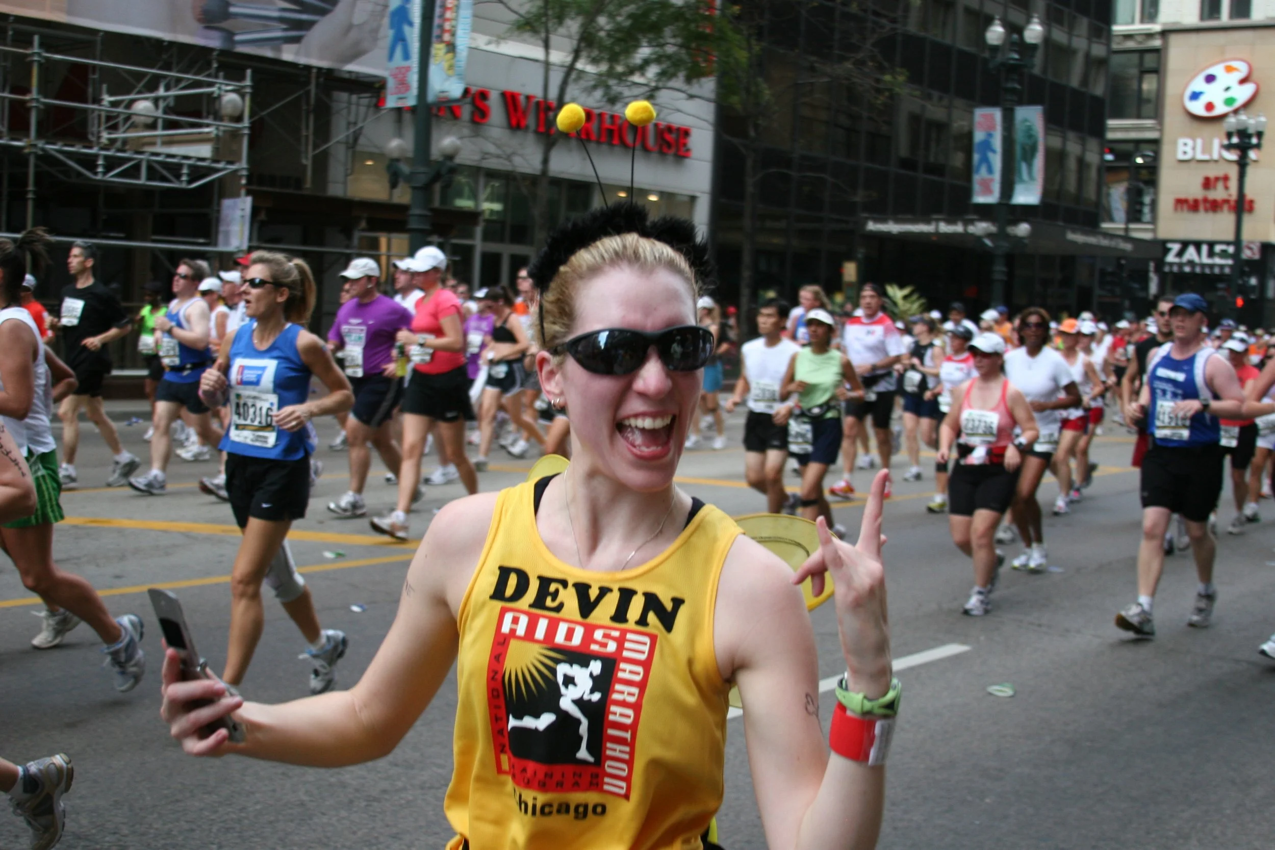 A smiling woman wearing sunglasses and a yellow tank top runs in a marathon on a city street, surrounded by other runners.