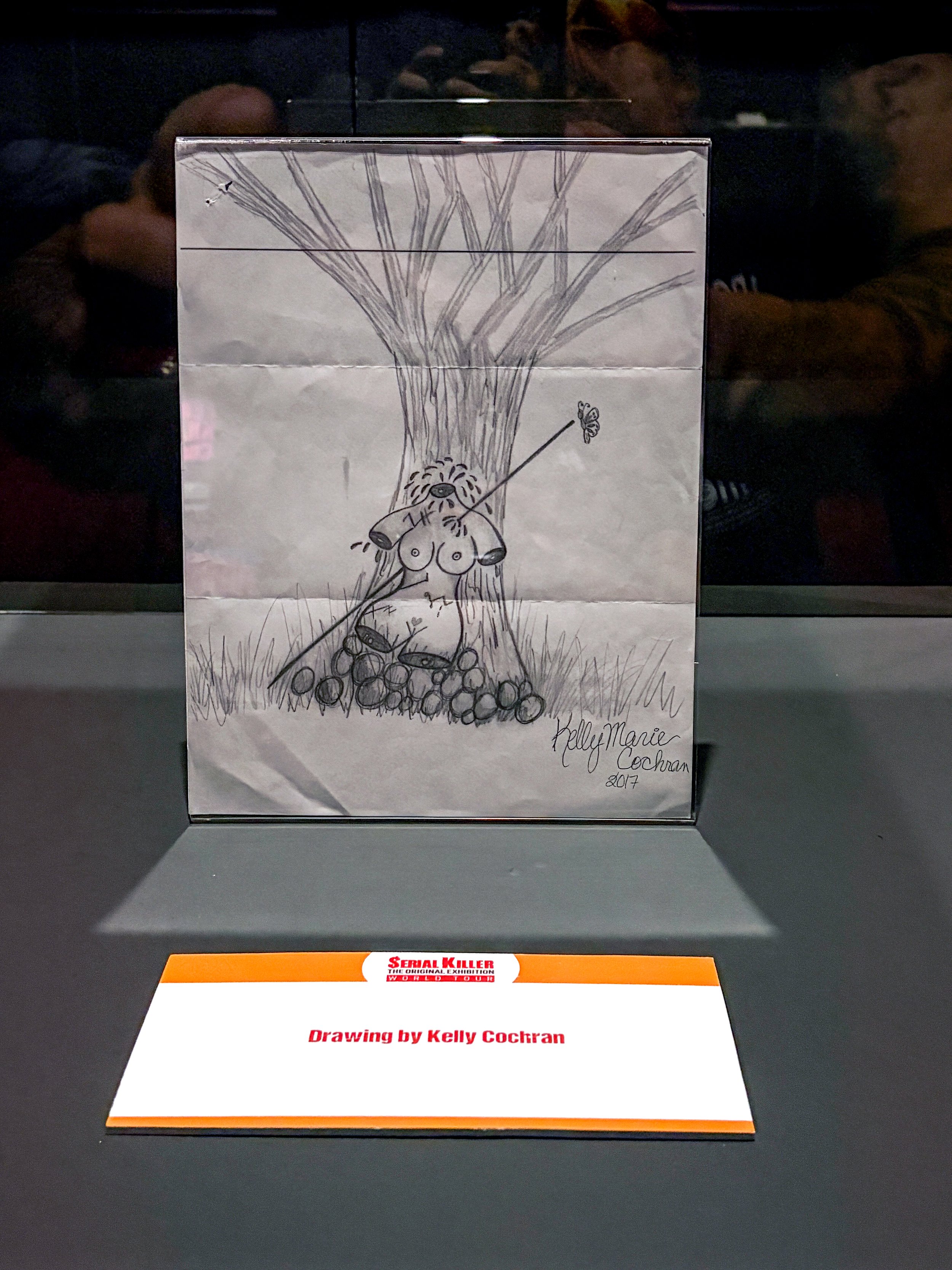 A black-and-white drawing showing a dog tied to a tree with a leash, with a butterfly flying nearby, and the artist's signature, Kelly Marie Cochran, 2017.