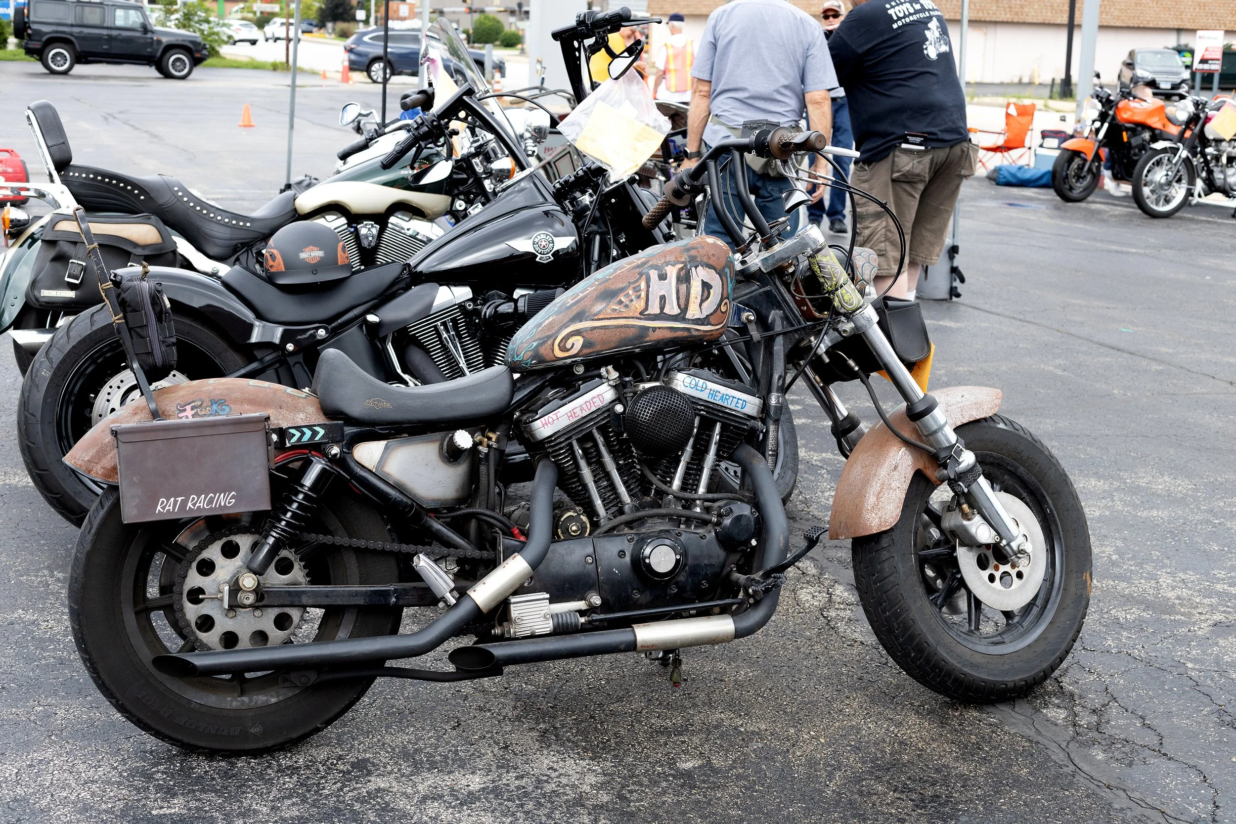Several motorcycles parked outdoors, with people standing and talking in the background. The motorcycles include a rusty, custom-painted bike with words 'HOT' and 'COLD' on the engine, and others with shiny black or chrome finishes. The scene appears