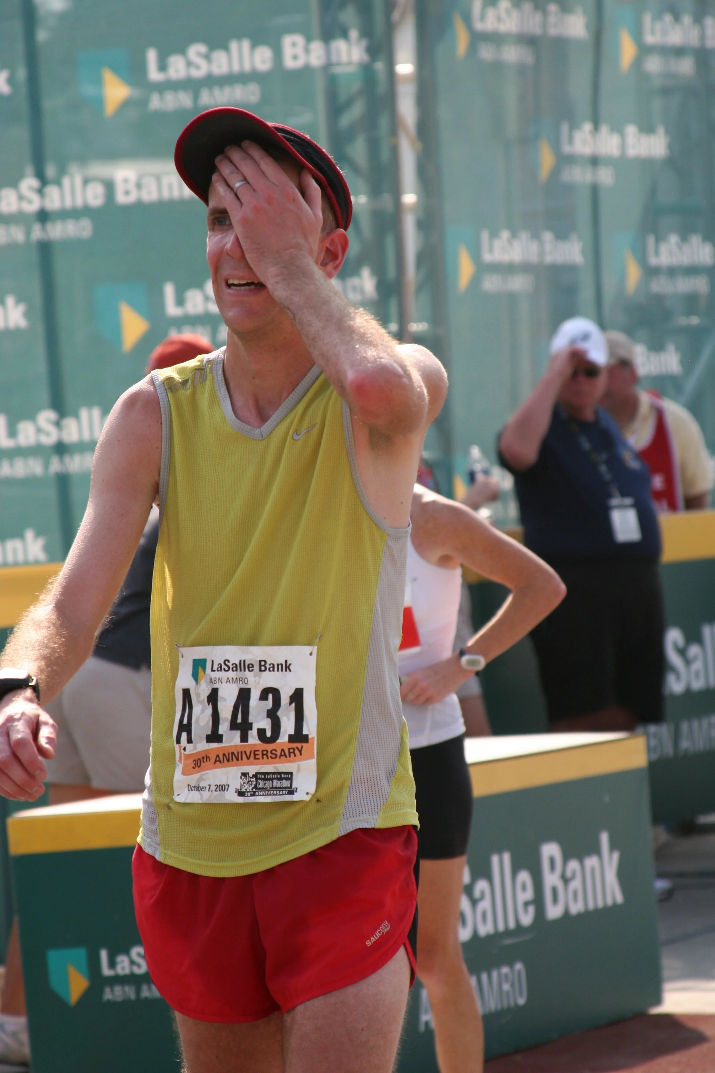 A male marathon runner wearing a yellow tank top and red shorts, touching his forehead, running past a LaSalle Bank sponsorship banner with other runners and officials in the background.