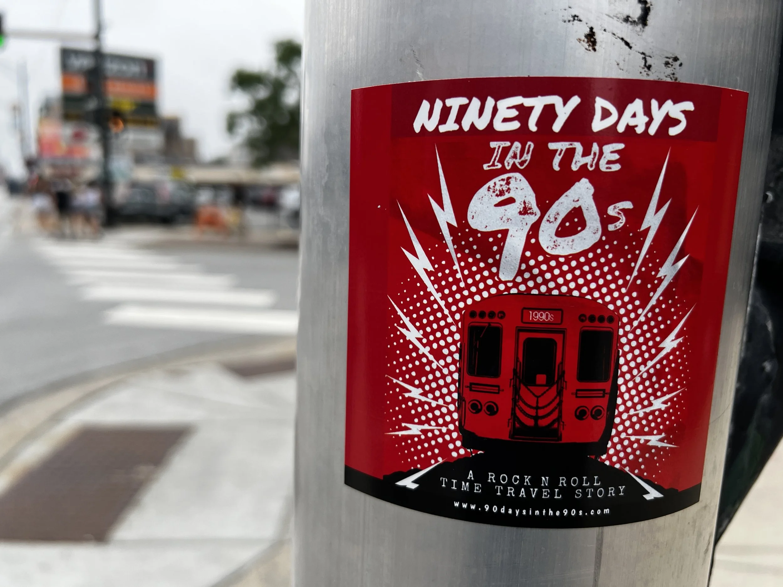 Red and black sticker on a metallic surface promoting a music album titled "Ninety Days in the 90s" with a graphic of a battery and the website www.90daysinthe90s.com
