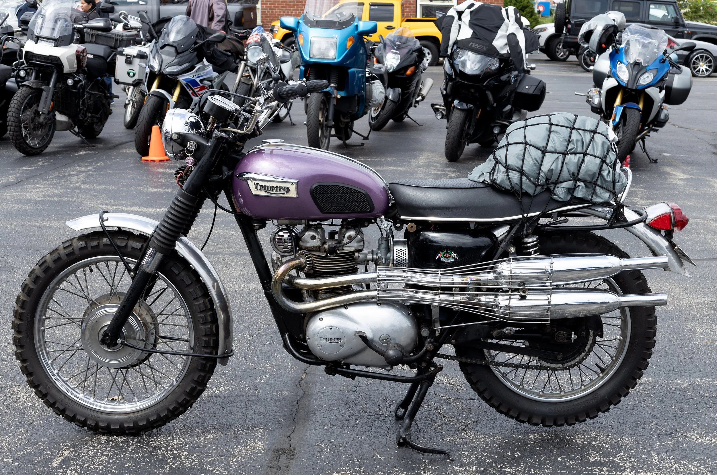 A purple Triumph motorcycle parked among several other motorcycles and scooters in a parking lot, with a brick building and cars in the background.
