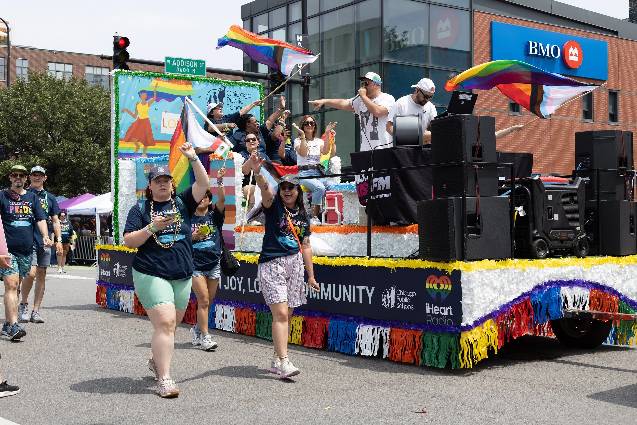 A parade float decorated with rainbow colors, featuring a Chicago Public Schools banner and iHeart Radio branding, surrounded by people celebrating during a Pride event on a city street.