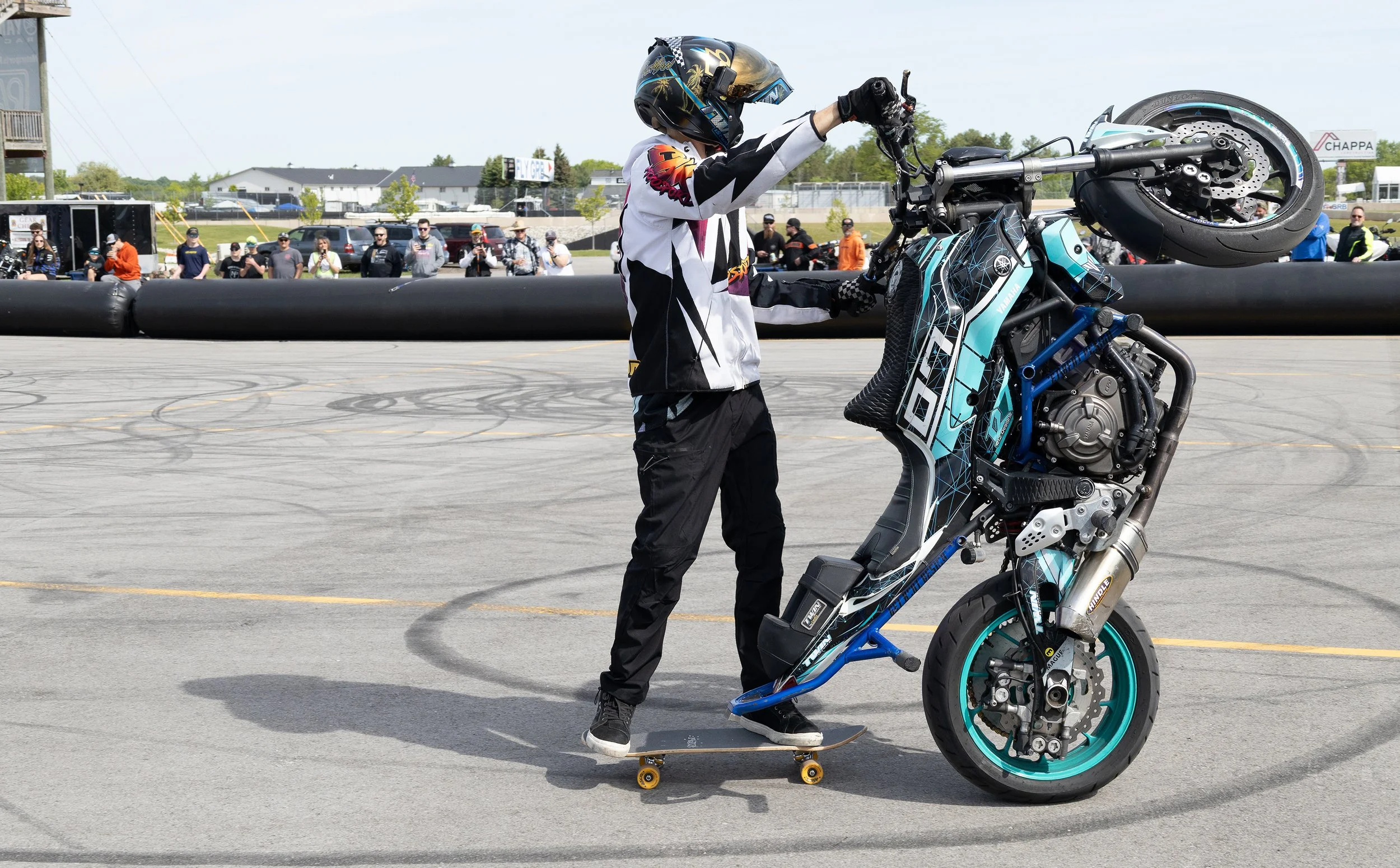 A person in a helmet and racing gear performs a wheelie stunt on a blue electric motorcycle at an outdoor event, with spectators watching in the background.