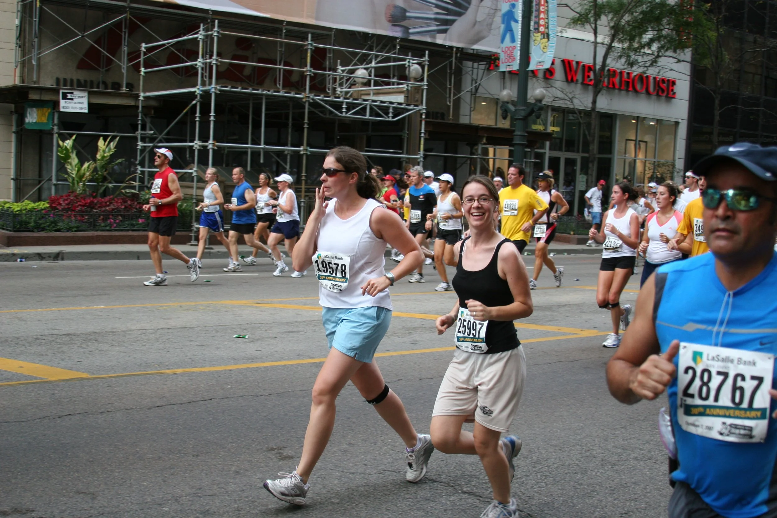 People participating in a marathon or running event on city streets, some smiling and wearing numbered bibs, with a background of a building, scaffolding, and trees.