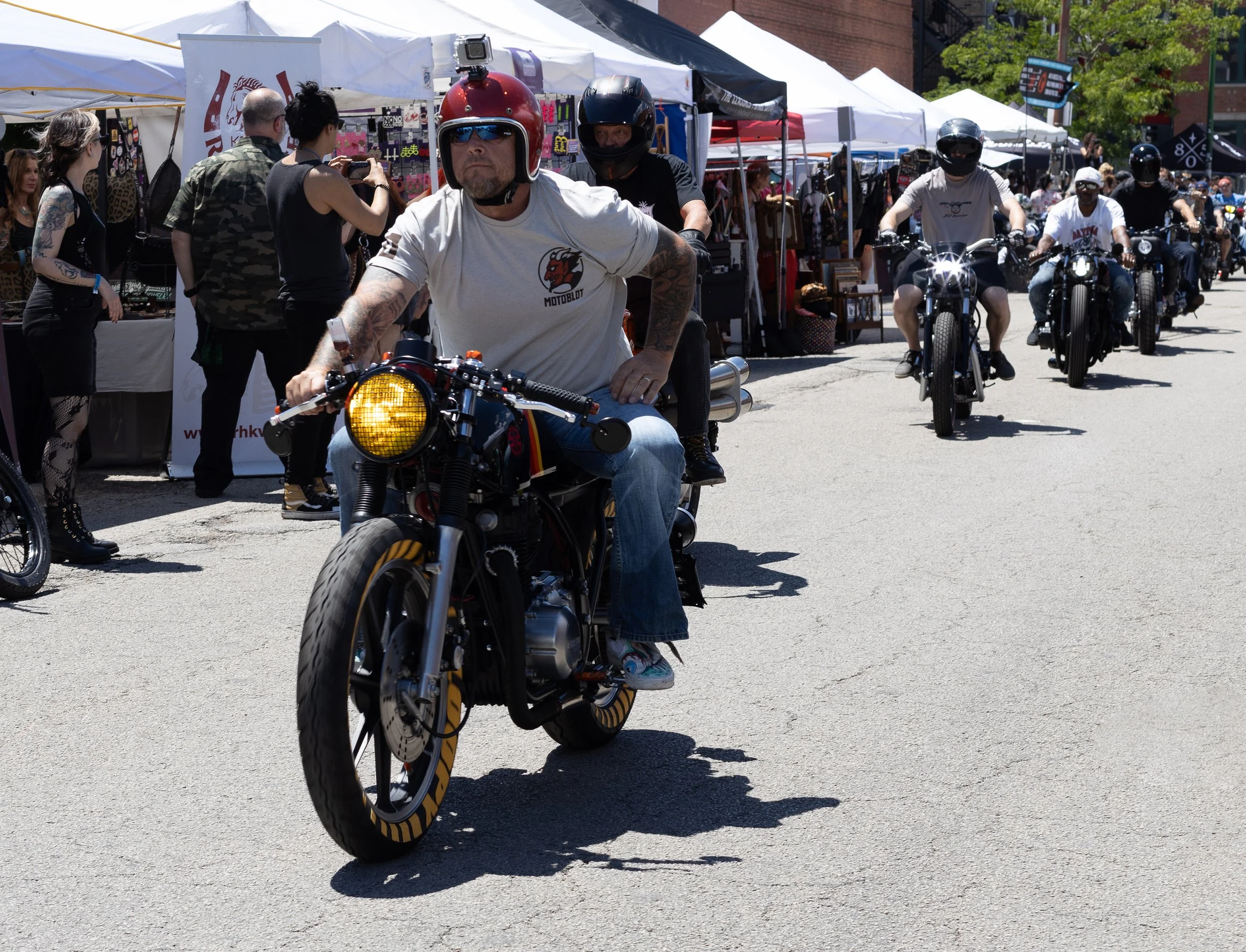 A group of motorcyclists riding in a parade or event, with market stalls and tents along the street, and people taking photos in the background.