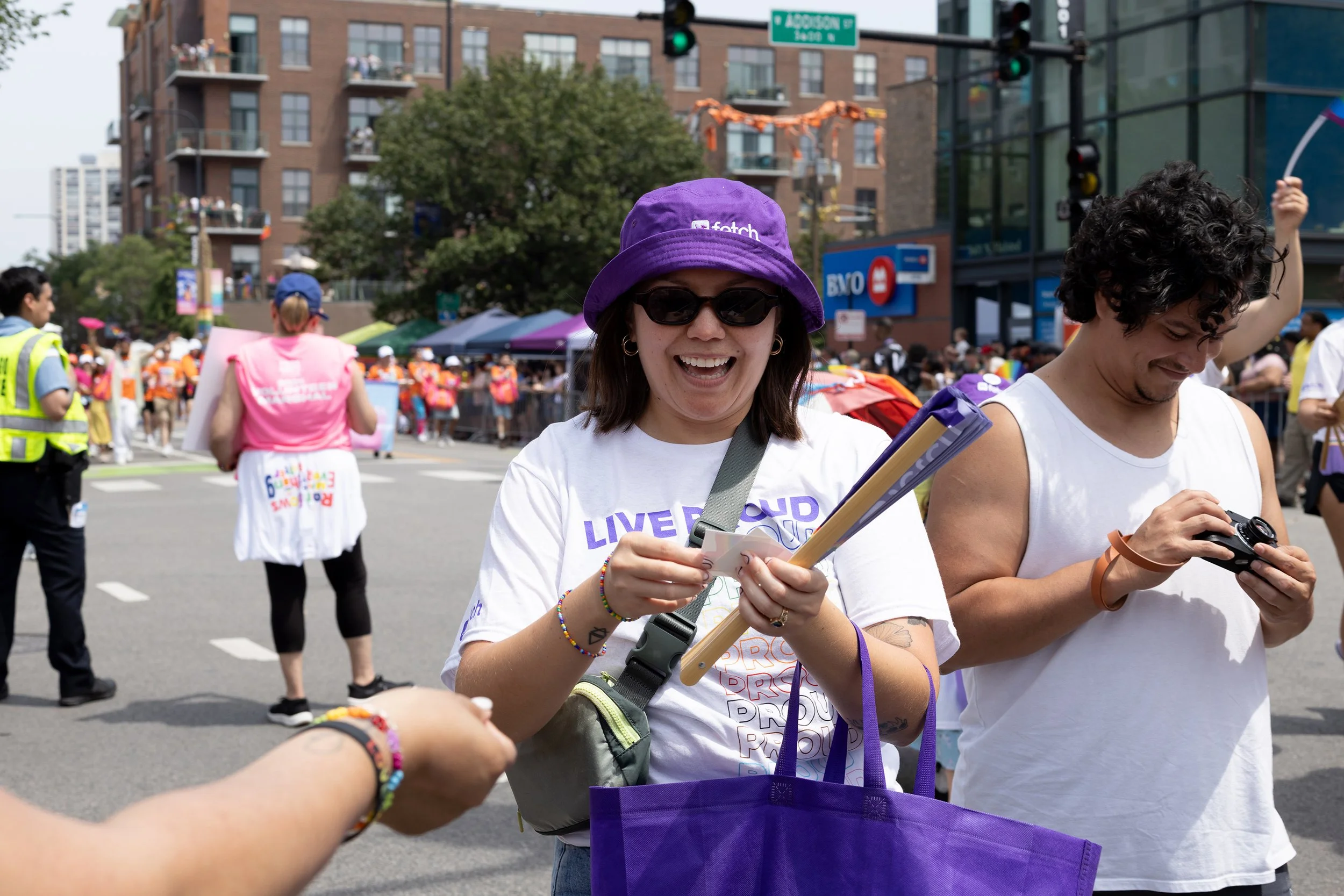 People participating in an outdoor event or parade in an urban setting, with buildings in the background, some holding signs or bags, and a woman smiling and wearing a purple hat and sunglasses.