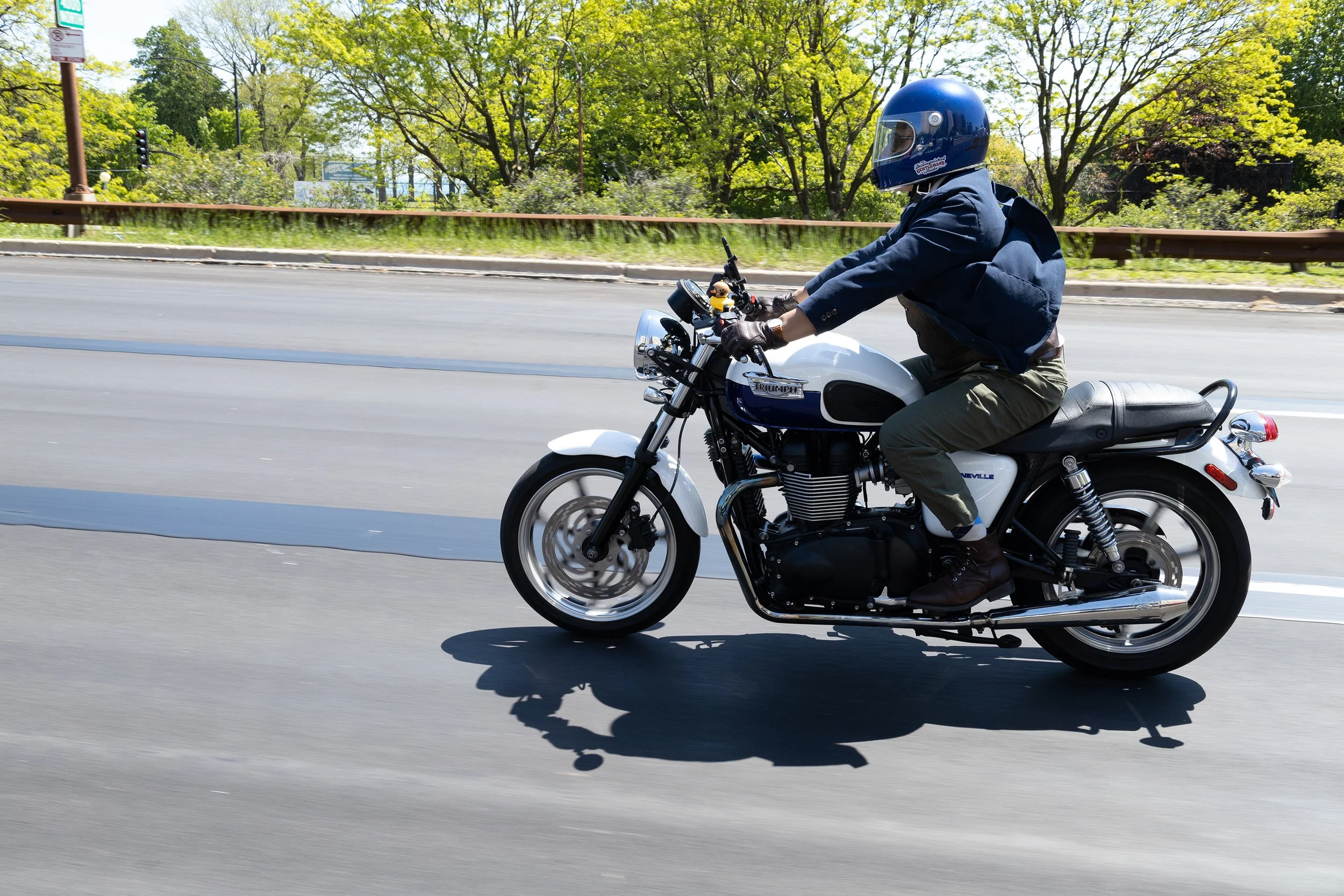 Person riding a Triumph motorcycle on a paved road with trees and greenery in the background, wearing a blue helmet, dark jacket, and green pants.
