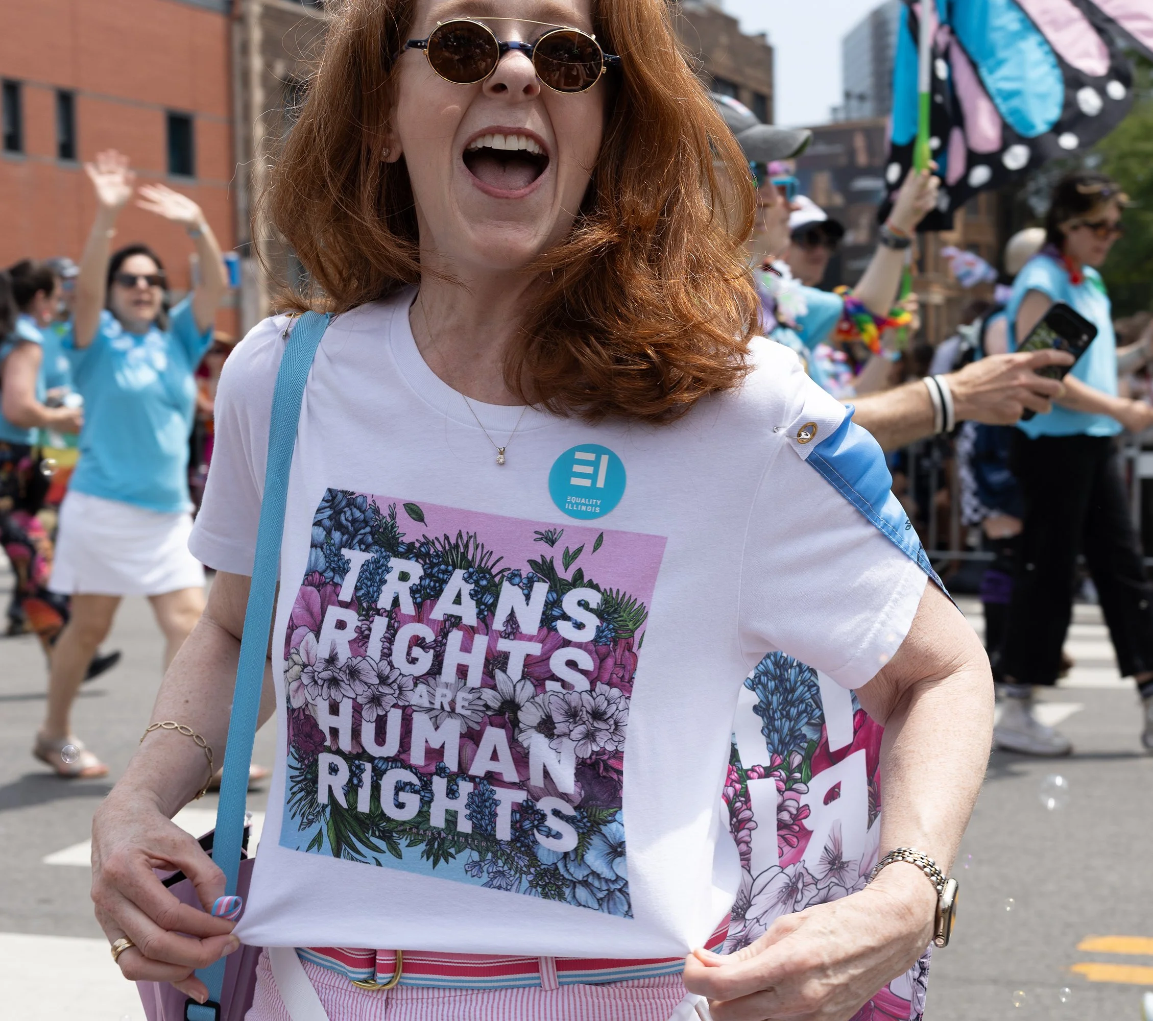 A woman with red hair wearing sunglasses, a white shirt with colorful floral print and the words "Trans Rights Are Human Rights" on it, participating in a public rally or march, with other people in the background, some wearing blue shirts and holdin