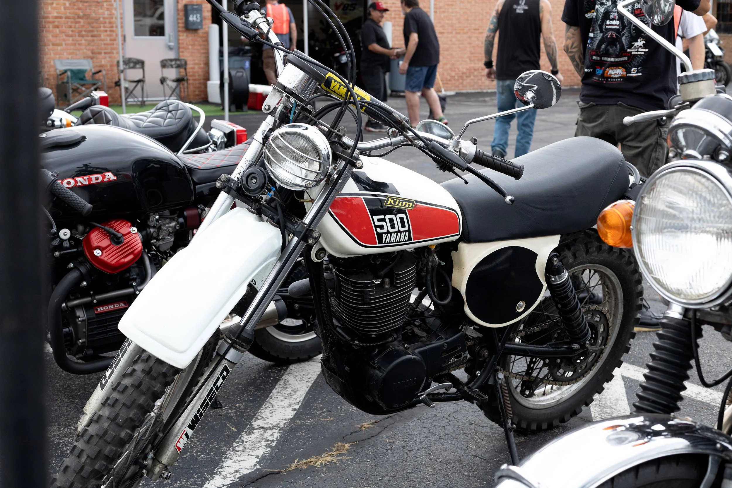 Close-up of a vintage Yamaha KLIM 500 motorcycle with a white fuel tank, surrounded by other motorcycles and people at a gathering or event.