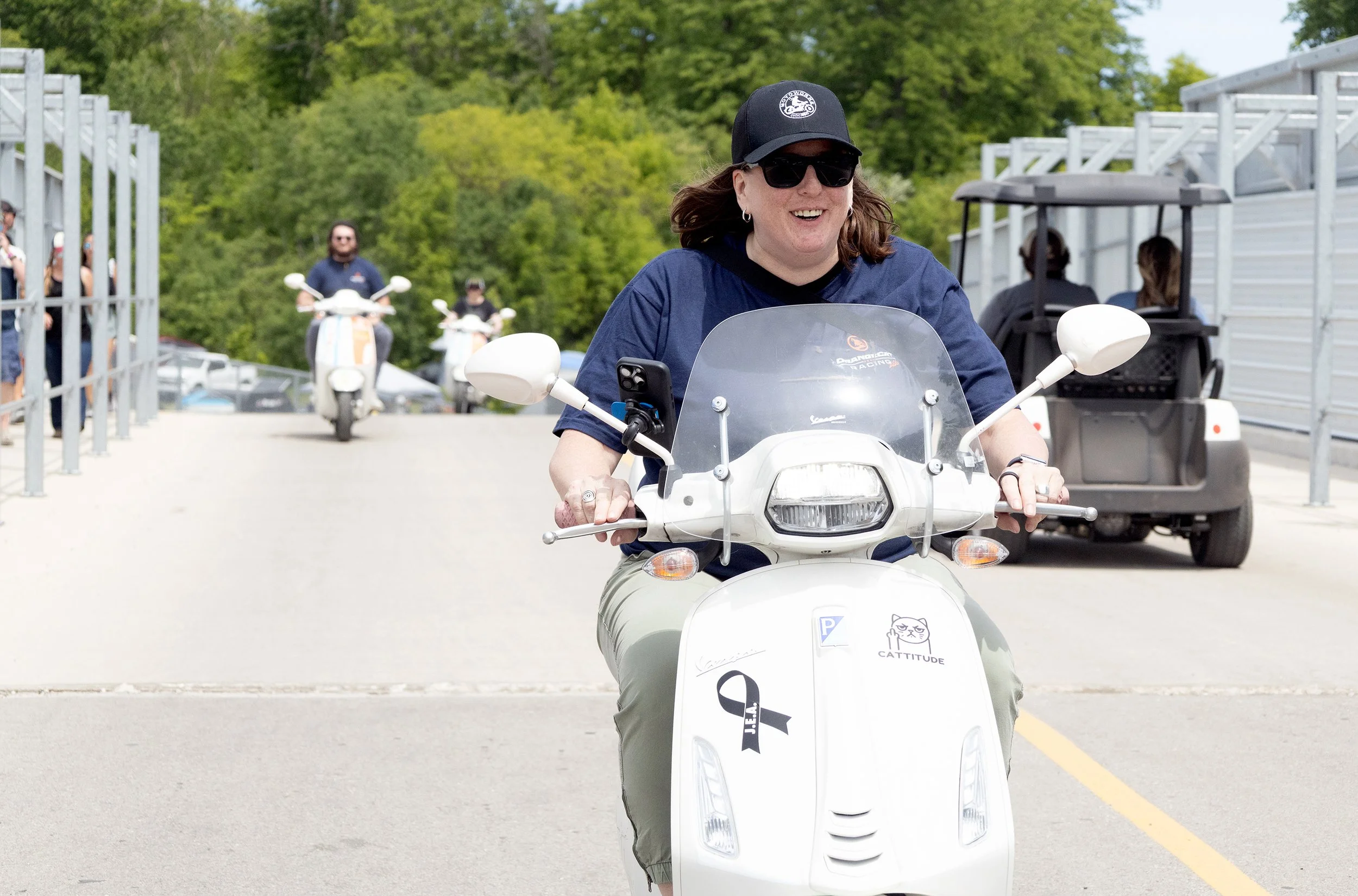 Woman riding a white scooter with a front windshield, wearing a black cap, sunglasses, and a blue shirt, smiling. In the background, there are trees, a golf cart with passengers, and a few other people riding scooters.