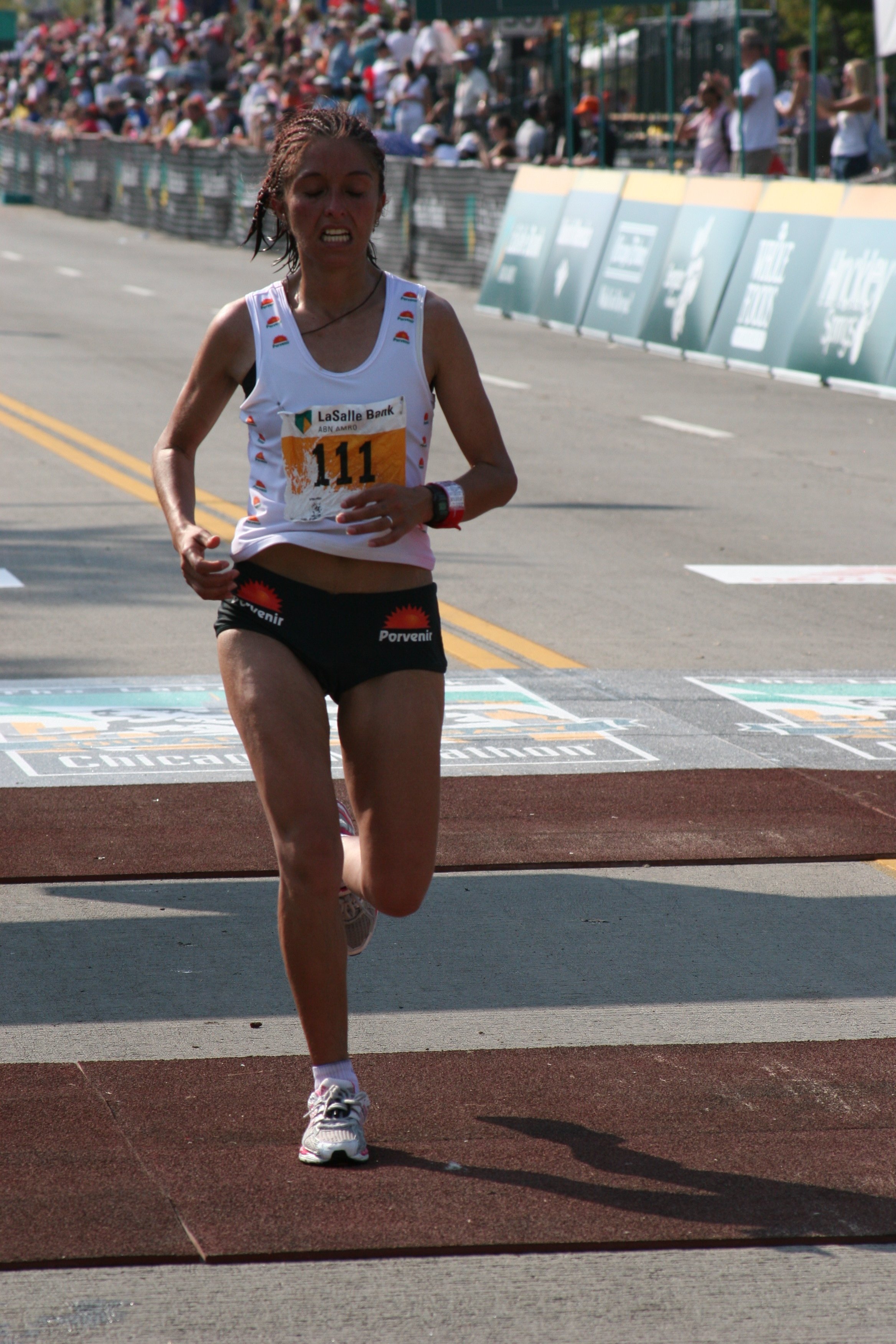 A female runner nearing the finish line in a race, with a crowd of spectators in the background.
