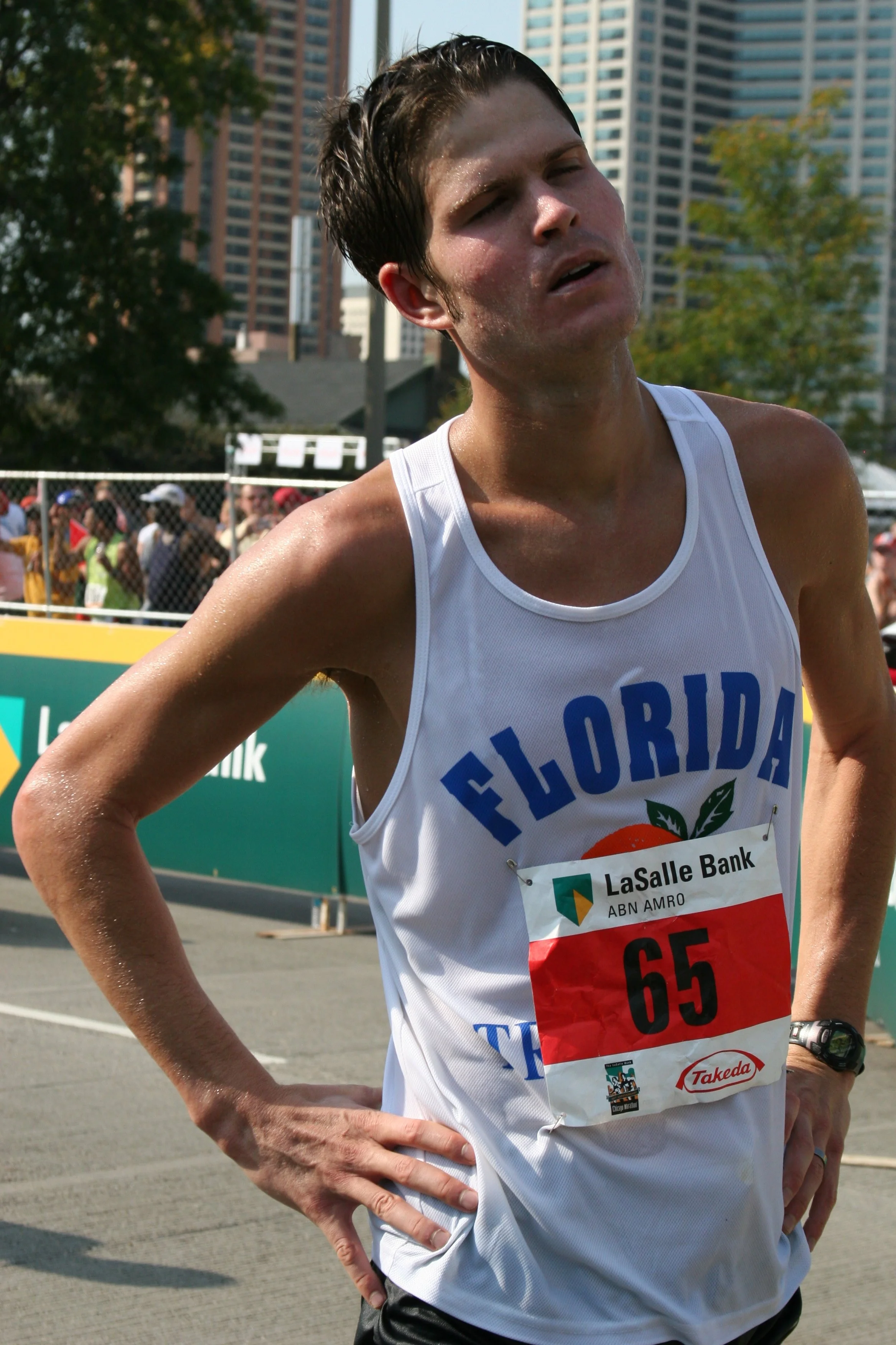 A male marathon runner wearing a white tank top and a red race bib with the number 65 standing outdoors in front of a crowd and tall buildings, with trees and a fence in the background.