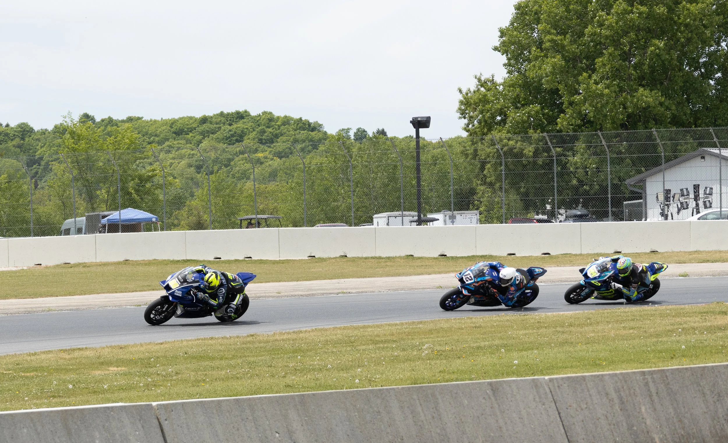 Motorcycle racers in a race leaning into a turn on a paved track. The background shows trees, a white fence, and some structures.