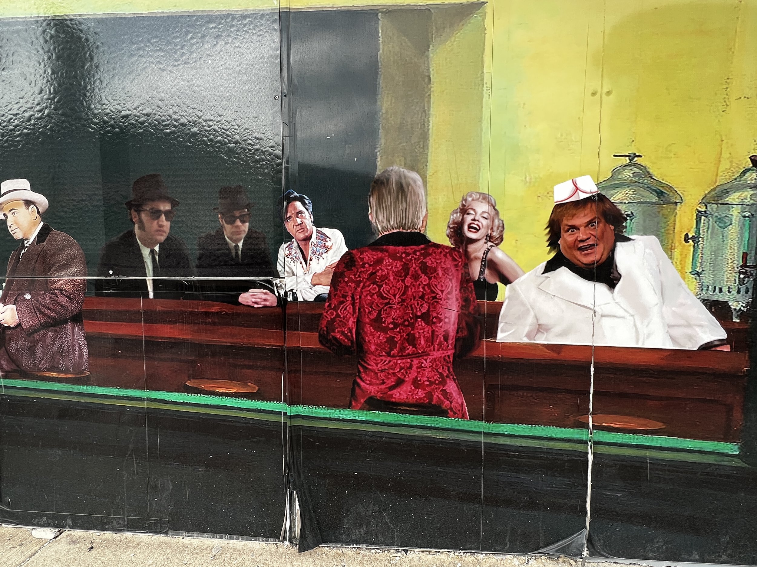 People in costume sitting at a bar with a painted landscape background, including trees and sky. One person is standing with their back to the camera, wearing a red velvet jacket. The others are engaged in conversation, some wearing hats and wigs.