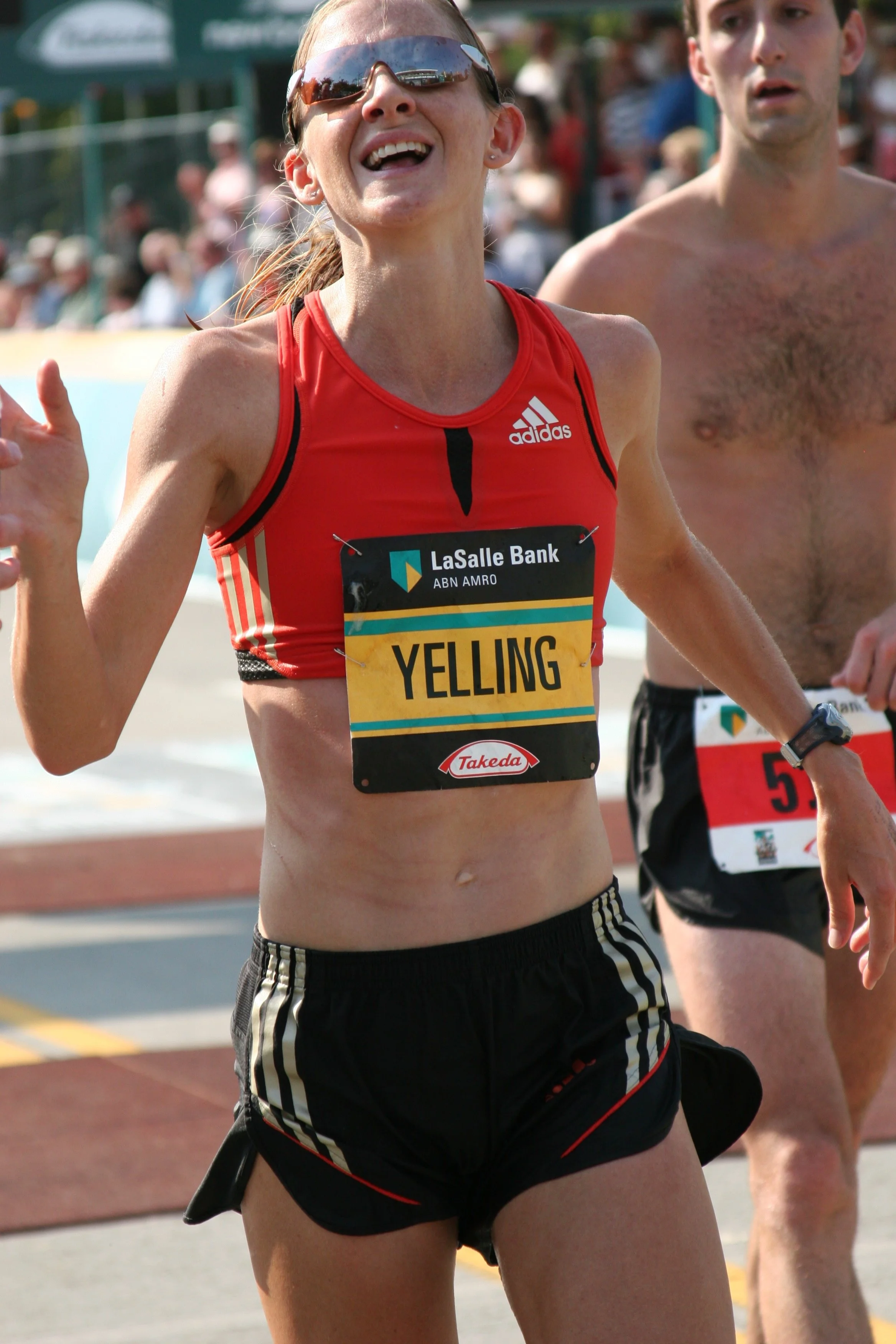 Close-up of a female marathon runner with long hair, sunglasses, wearing a red Adidas top and black shorts, smiling and running during a race.