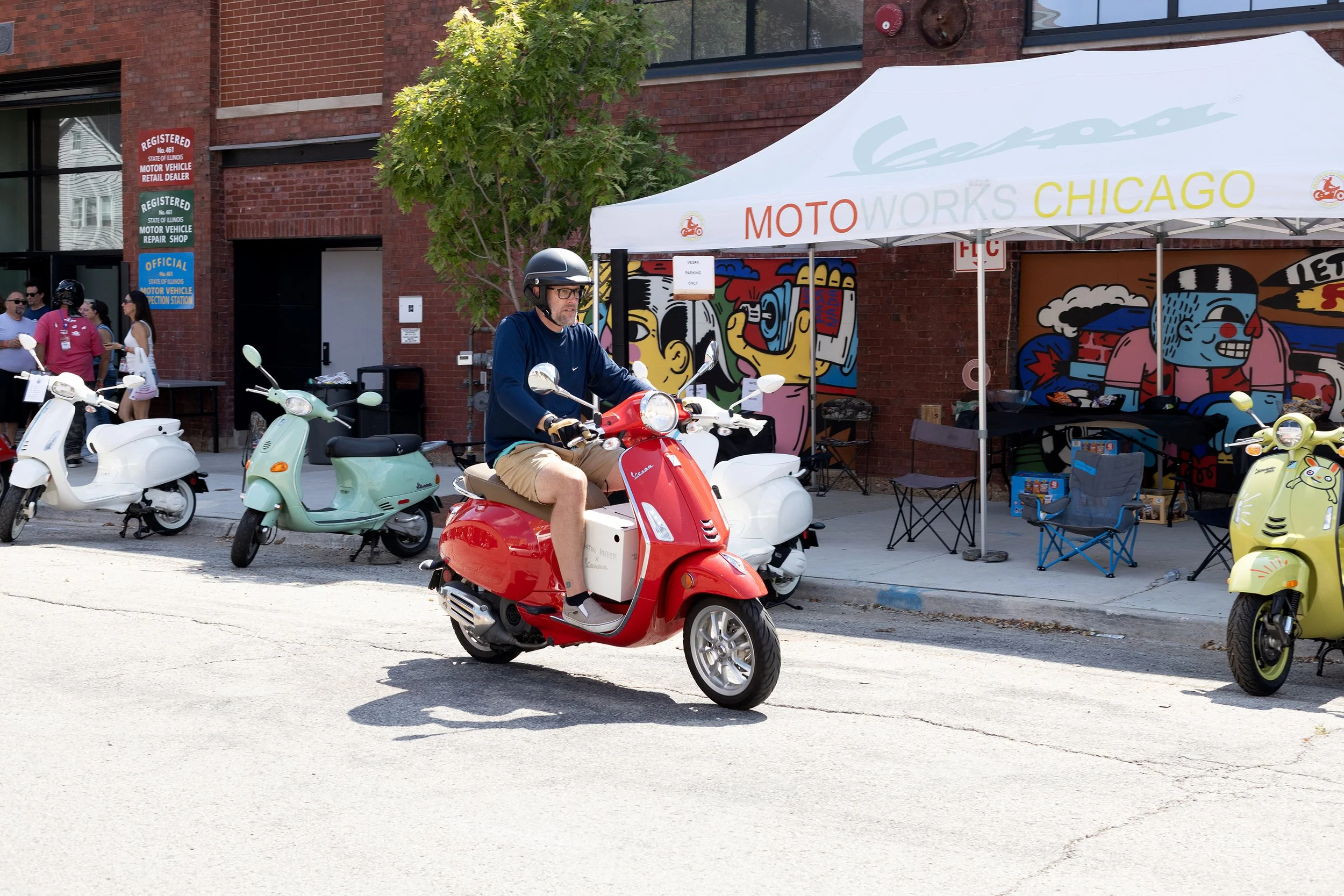 A man wearing a helmet and glasses riding a red scooter on the street near a motorcycle event booth with colorful graffiti art on the wall behind.