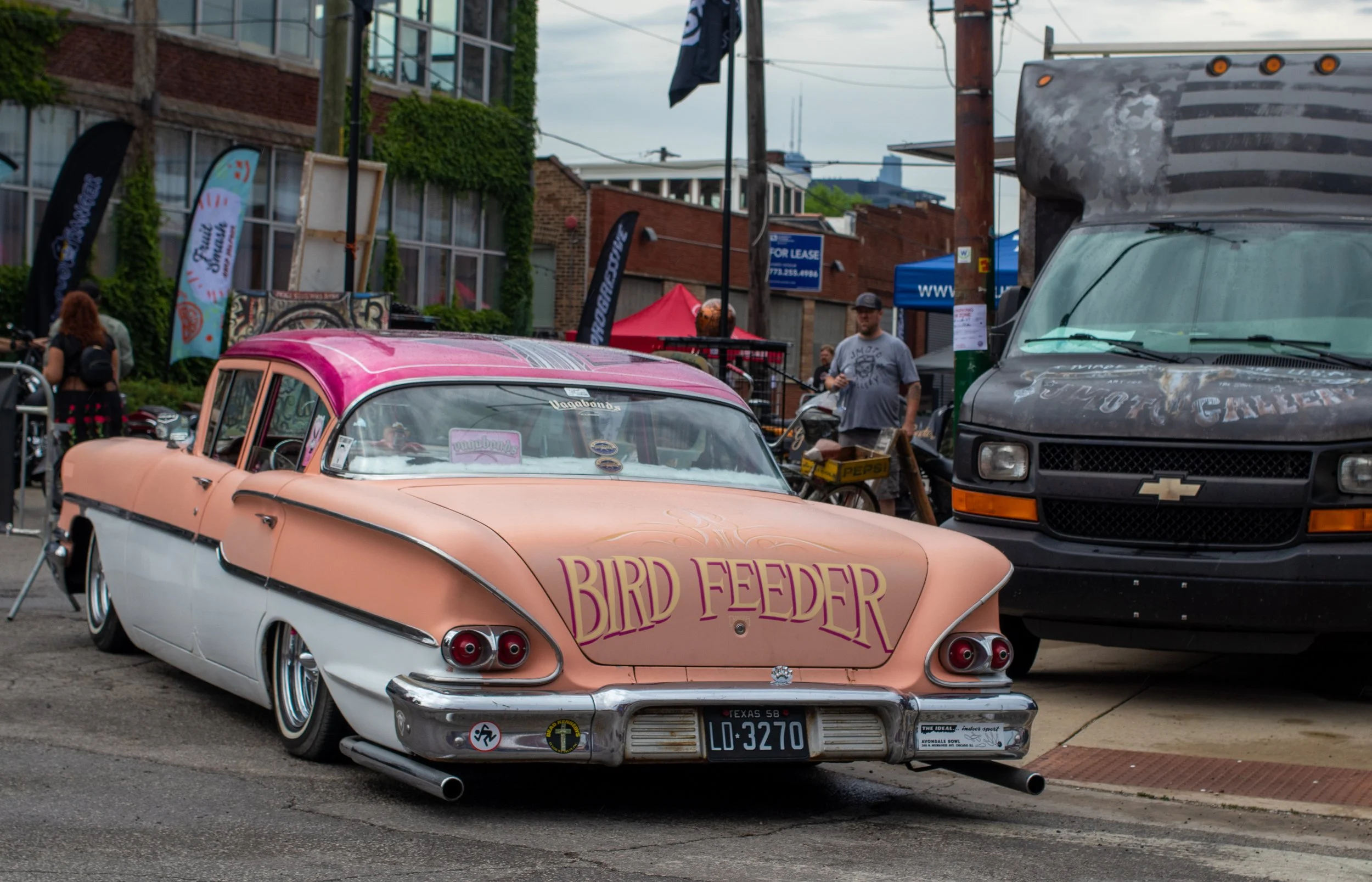 A vintage pink and white Cadillac with the words 'Bird Feeder' written on its side, parked on a street at an outdoor event with people, tents, flags, and a large black van nearby.