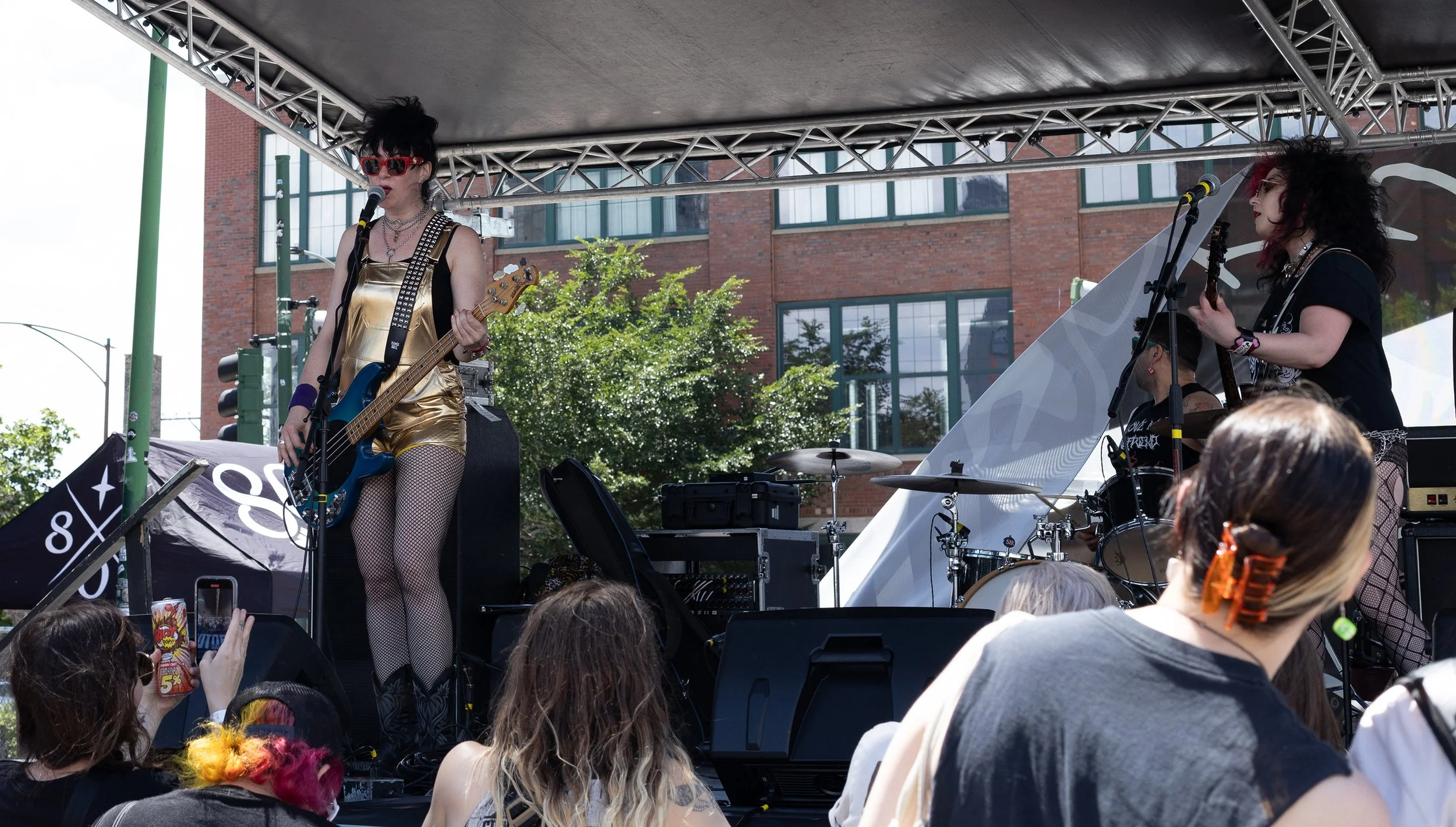 A female musician with black hair and red sunglasses playing bass guitar on an outdoor stage, with another female musician singing and playing guitar, surrounded by audience members and equipment, with buildings and trees in the background.