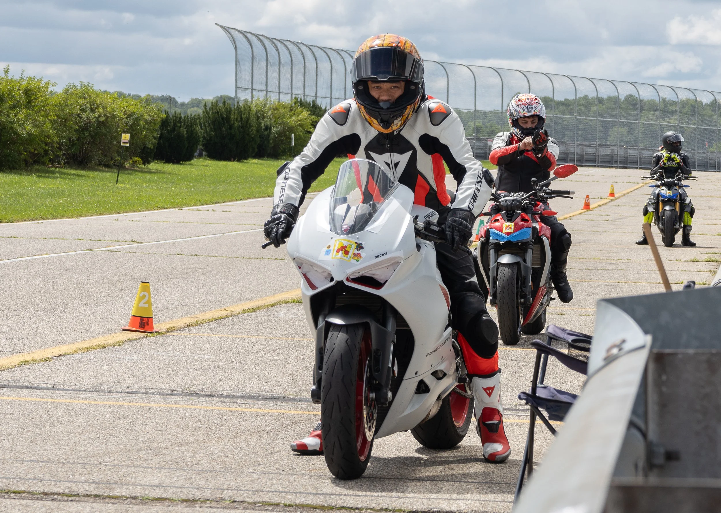 Motorcyclists participating in a riding event on an outdoor track, with cones and markers, under a partly cloudy sky.