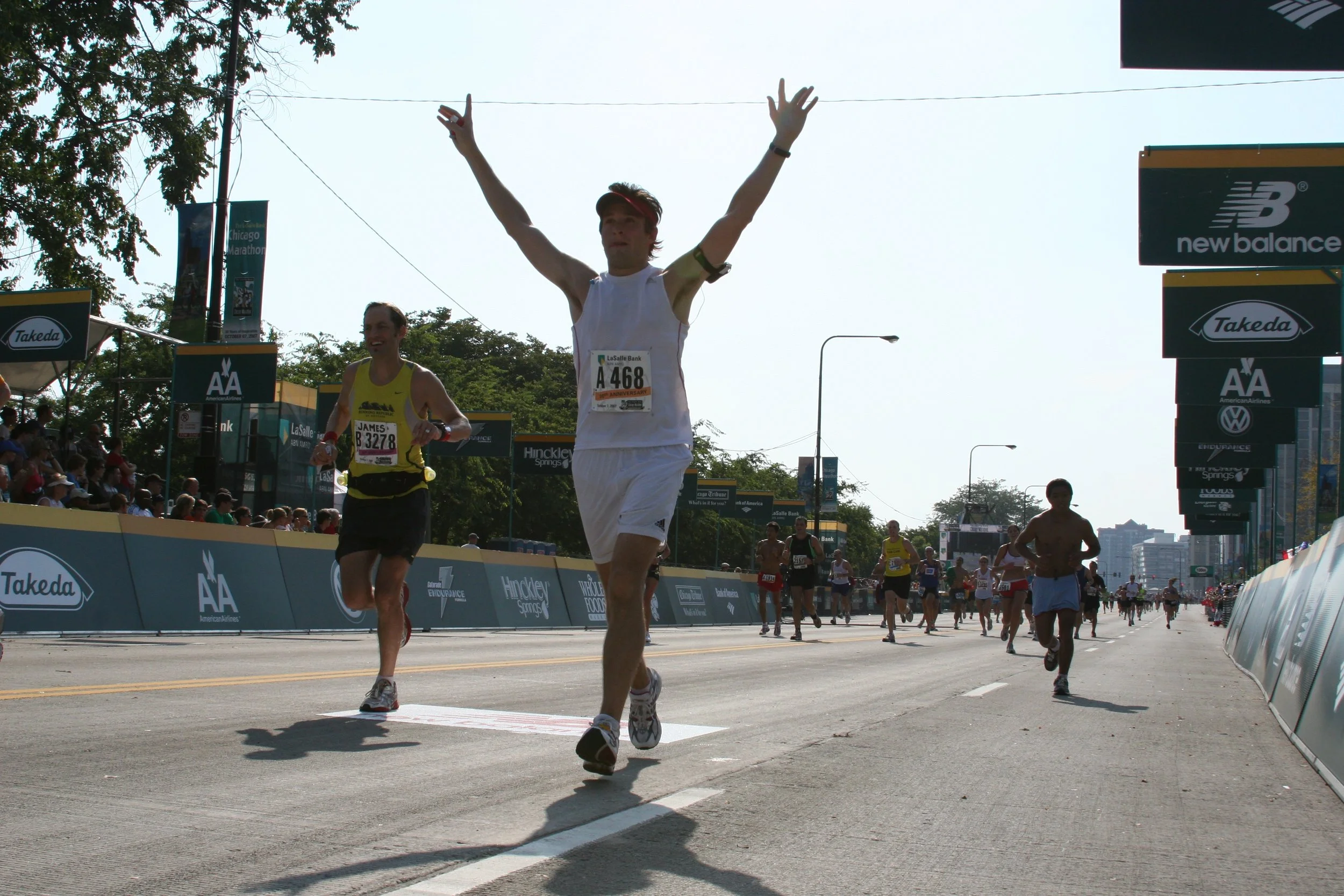 Marathon runners crossing the finish line at a road race in an urban area, with spectators on the sides and banners overhead.