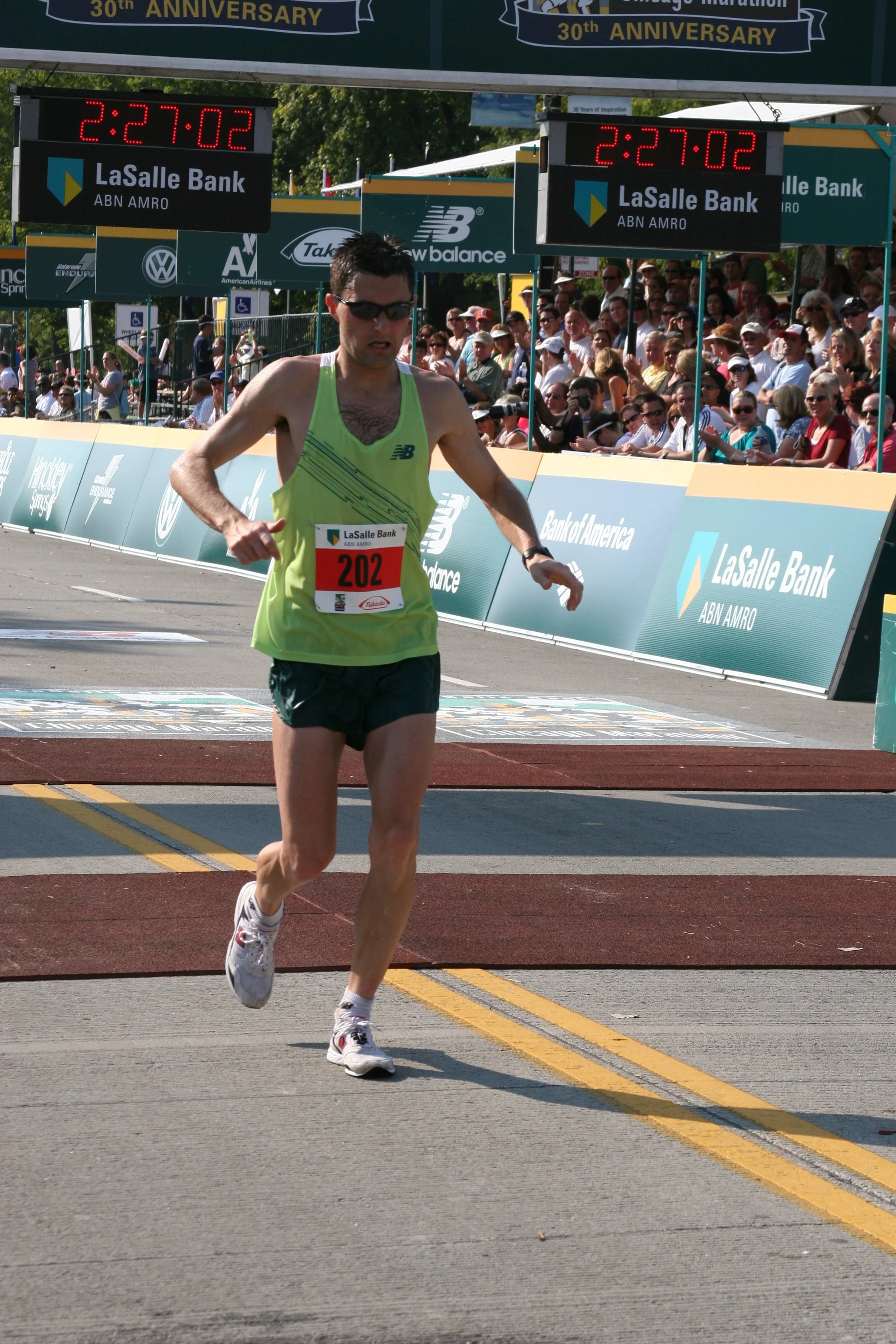 A male runner with bib number 202 approaching the finish line at a marathon race, with a crowd of spectators behind barriers and digital clocks displaying 2:27:02.