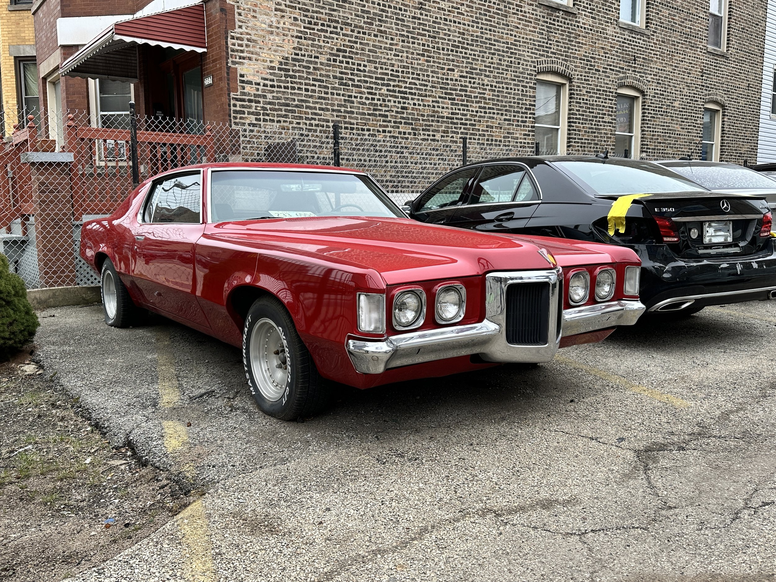 A red vintage car parked next to a black luxury Mercedes-Benz E350 with yellow tape on the trunk, in an urban setting with brick buildings and a chain-link fence in the background.