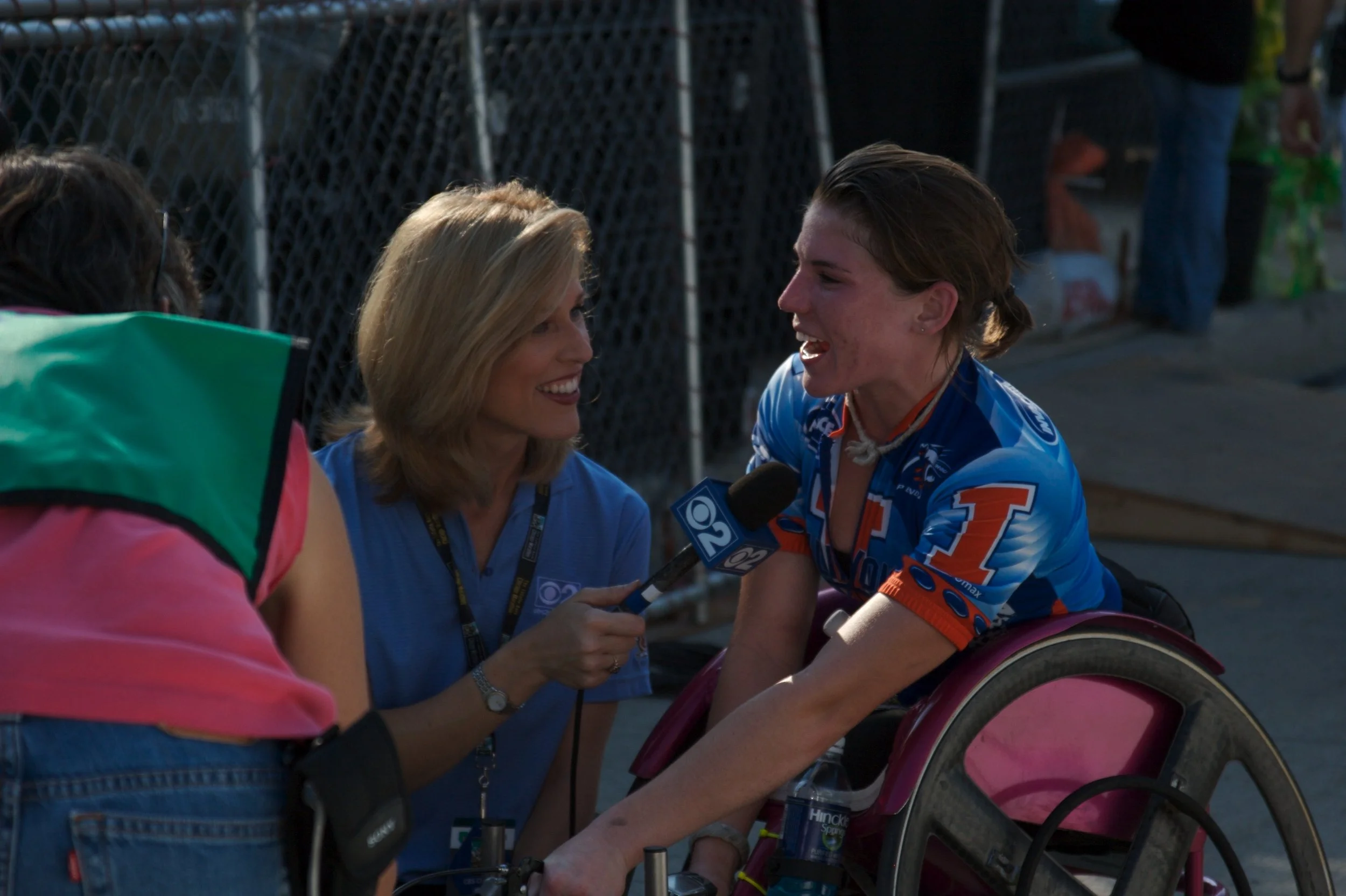 A woman in a racing suit being interviewed by a reporter holding a microphone. The woman appears to be a cyclist or athlete with a wheelchair. The scene seems to be outdoors, near a fence, possibly after a race or event.