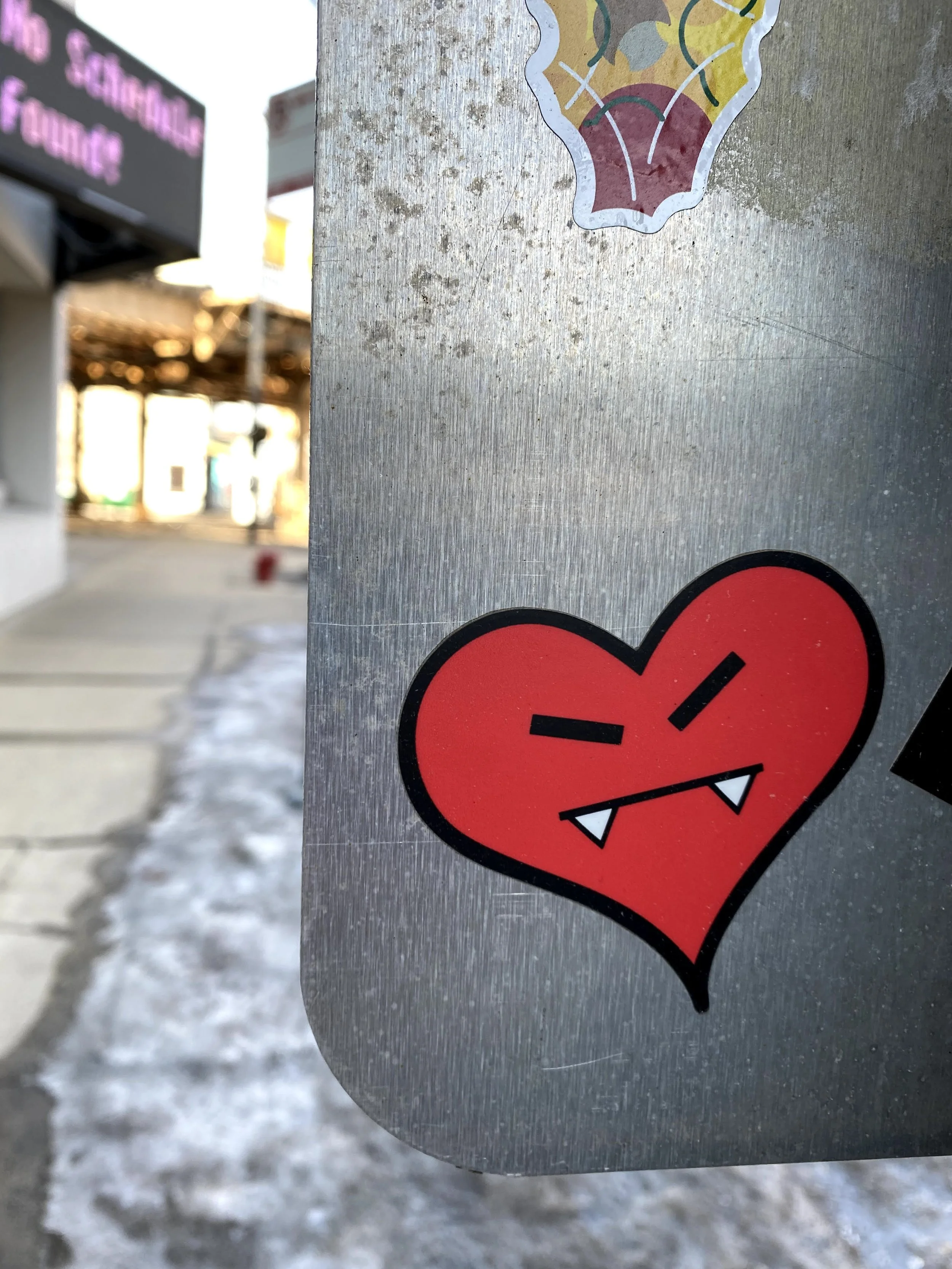 Close-up of a damaged red heart-shaped sticker with fangs on a metallic surface in a winter outdoor setting, with store signs and a sidewalk in the blurry background.