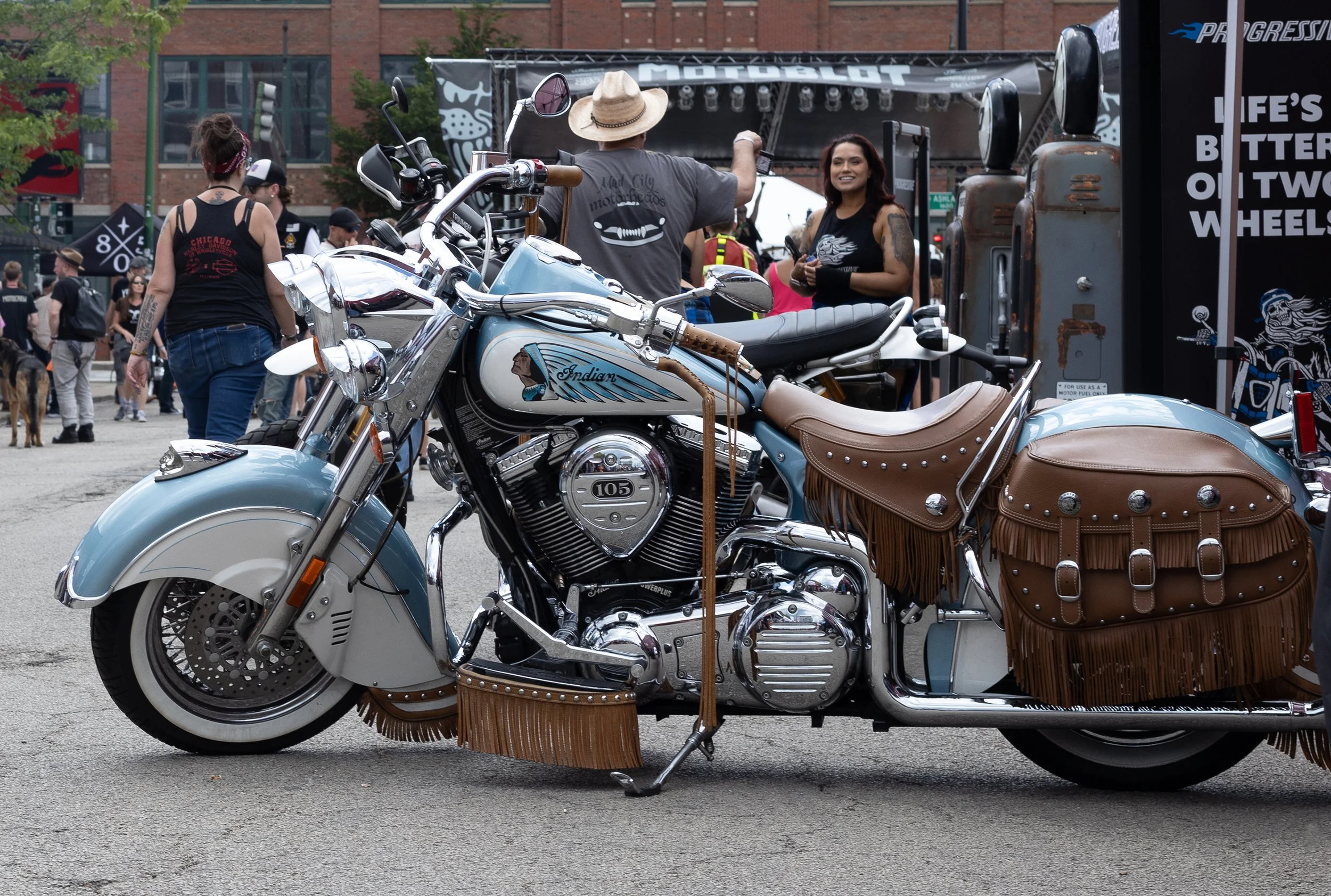 A vintage light blue Harley Davidson motorcycle with leather saddlebags and fringe detailing, parked at an outdoor event. In the background, people are gathered, some wearing black shirts, with a stage and banners visible.