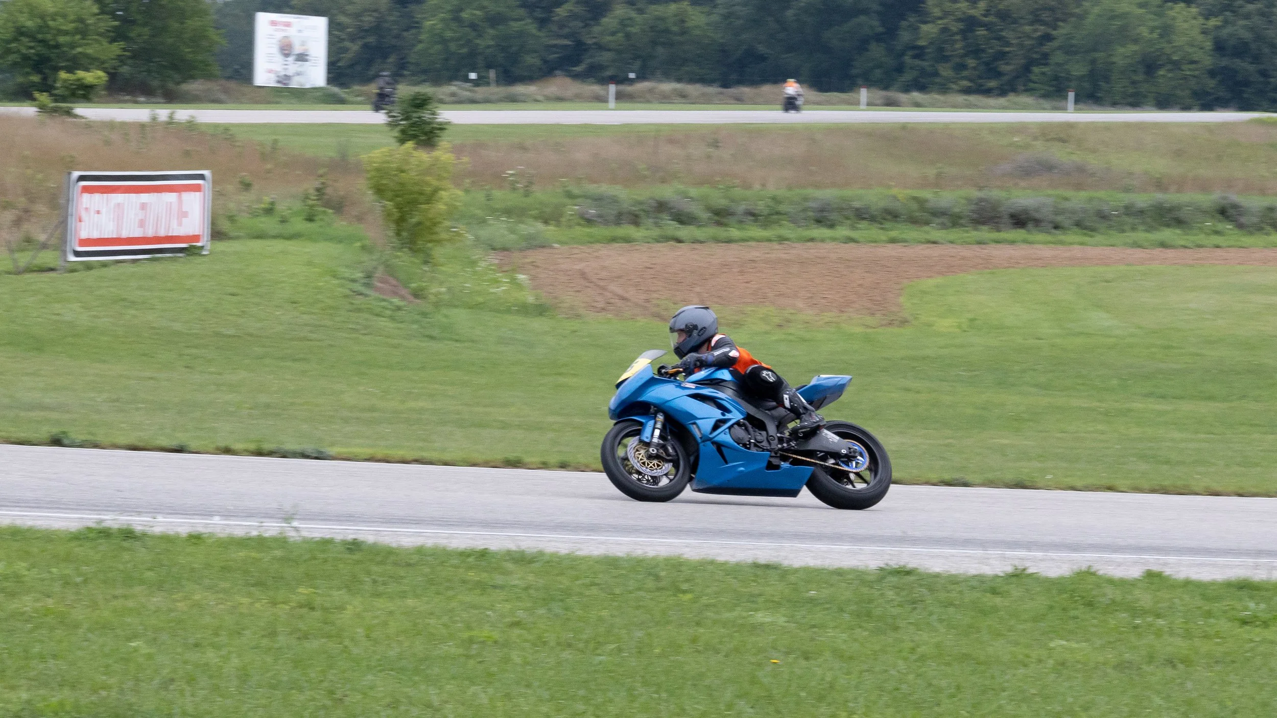 A motorcyclist in a black helmet and orange jacket riding a blue sport bike on a paved road. Green grass and trees are in the background, along with some signs and a person riding a bicycle.