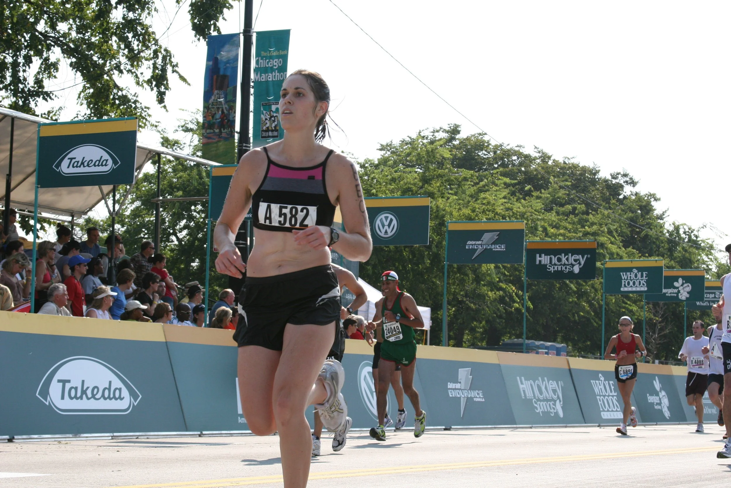 A female marathon runner with bib number A 582 running in a race, with other runners and an audience visible behind her at the Chicago Marathon, during daytime with trees and sponsor banners in the background.