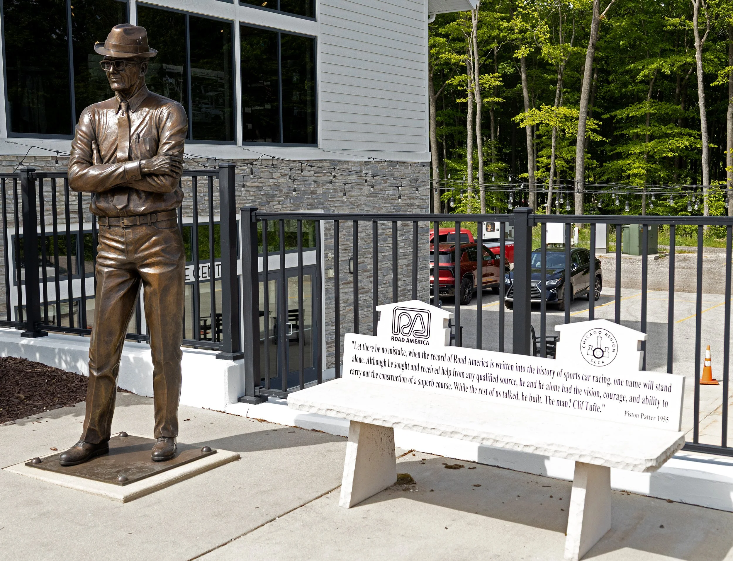 Bronze statue of a man wearing glasses, a hat, a long-sleeved shirt, and pants, standing with arms crossed in front of a fence. Behind the statue, there is a white building with large windows, a stone foundation, string lights, parked cars, and a woo
