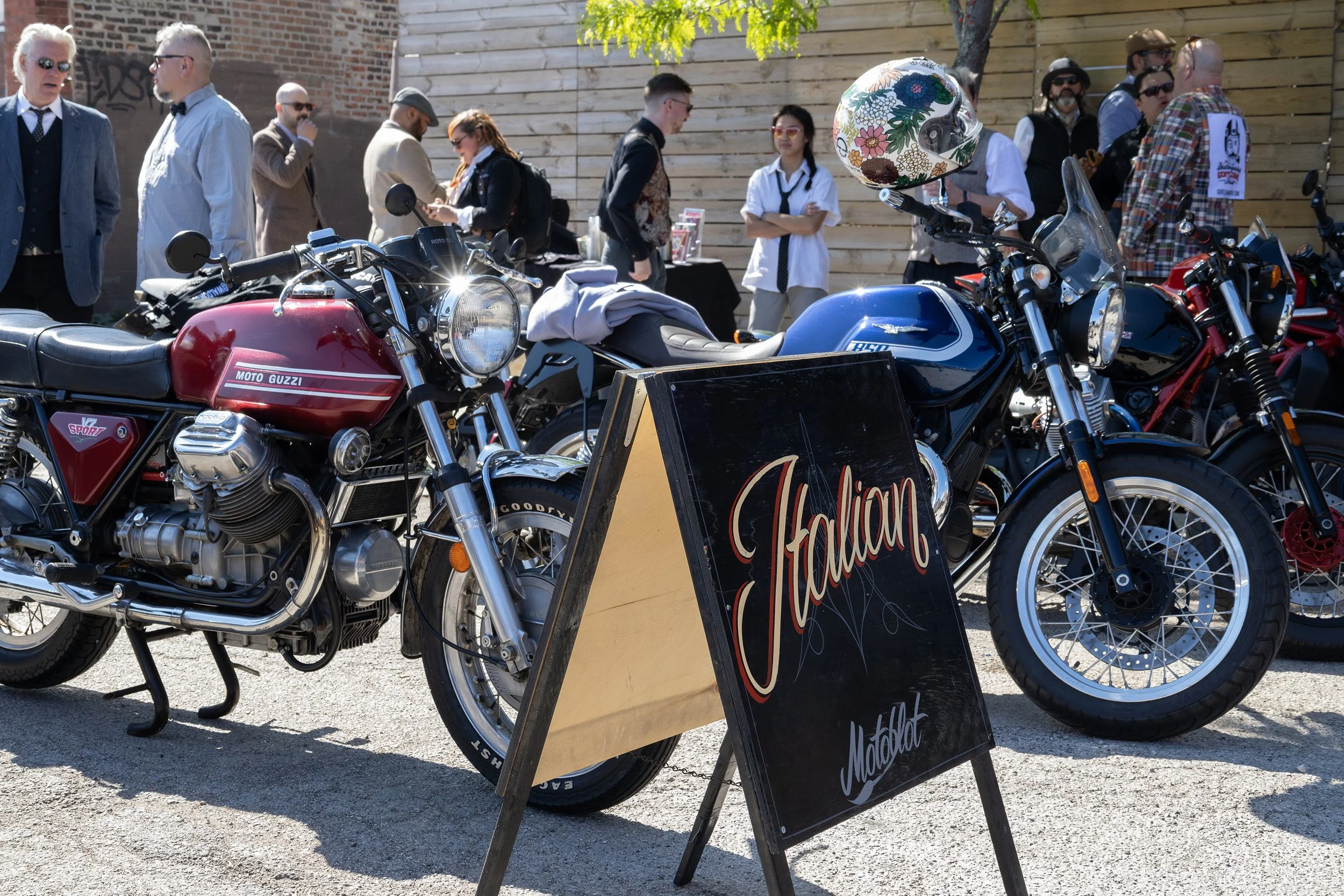 Multiple vintage motorcycles parked in front of a wooden wall at an outdoor event with people conversing. A sign with the word "California" is visible in the foreground.