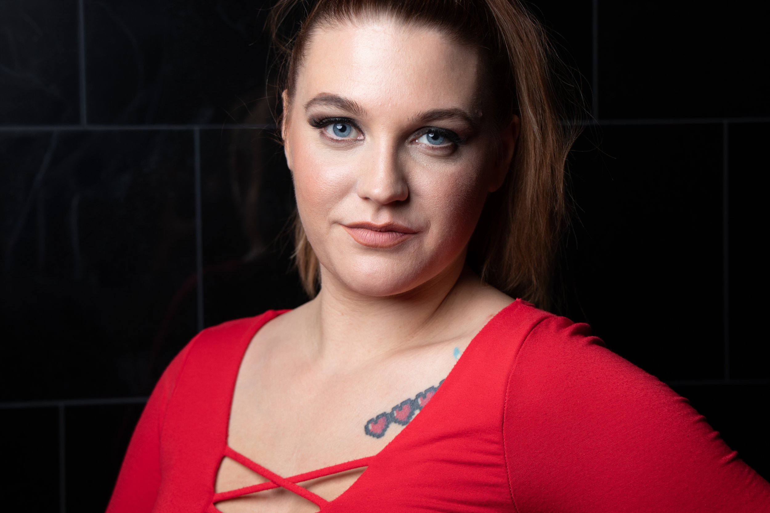 A woman with red hair and blue eyes wearing a red dress, standing against a black tiled wall.