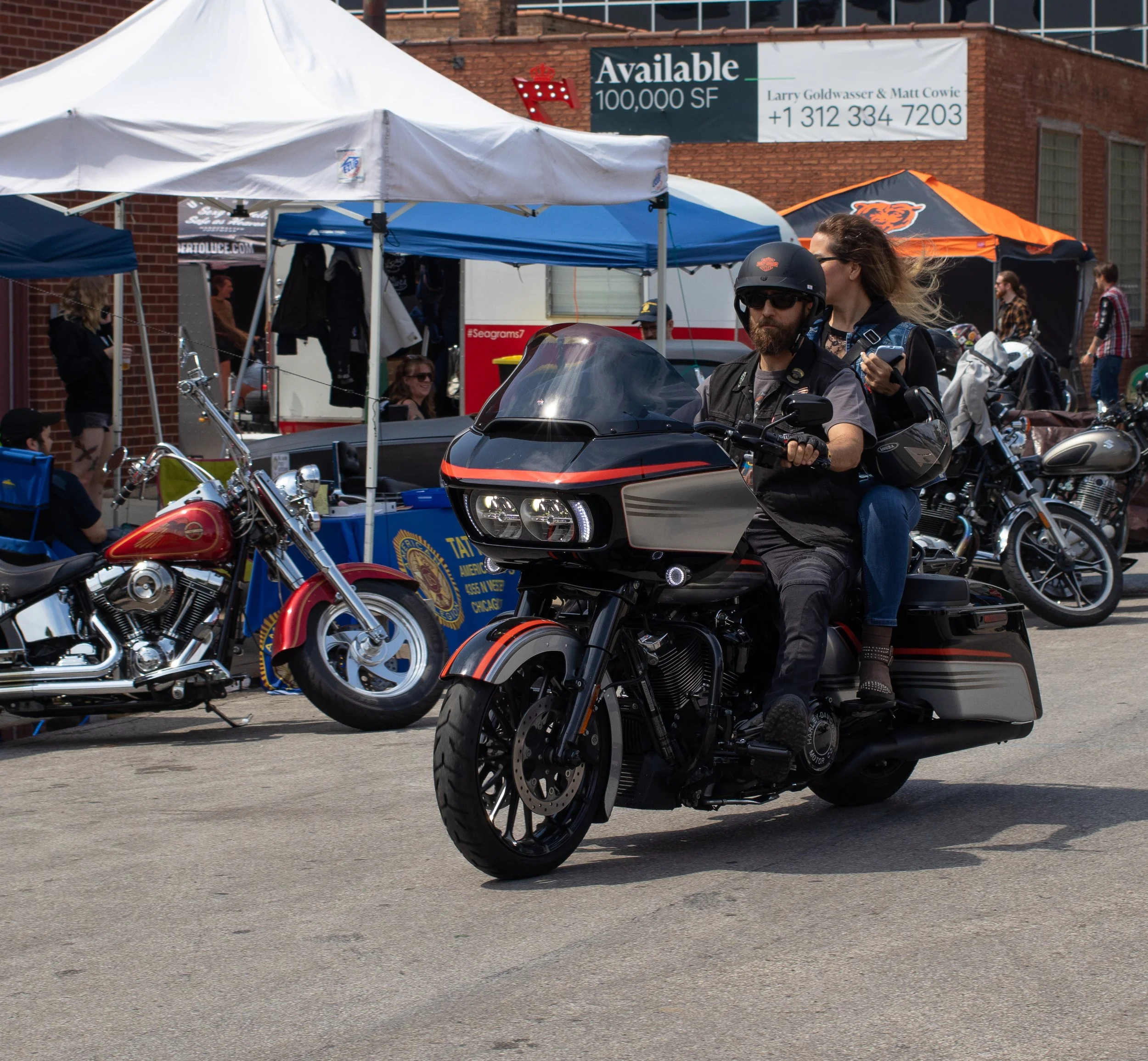 A man with long hair and a beard riding a black motorcycle with orange accents, wearing sunglasses, a helmet, and a vest. There are tents and people in the background, along with a building and a billboard.