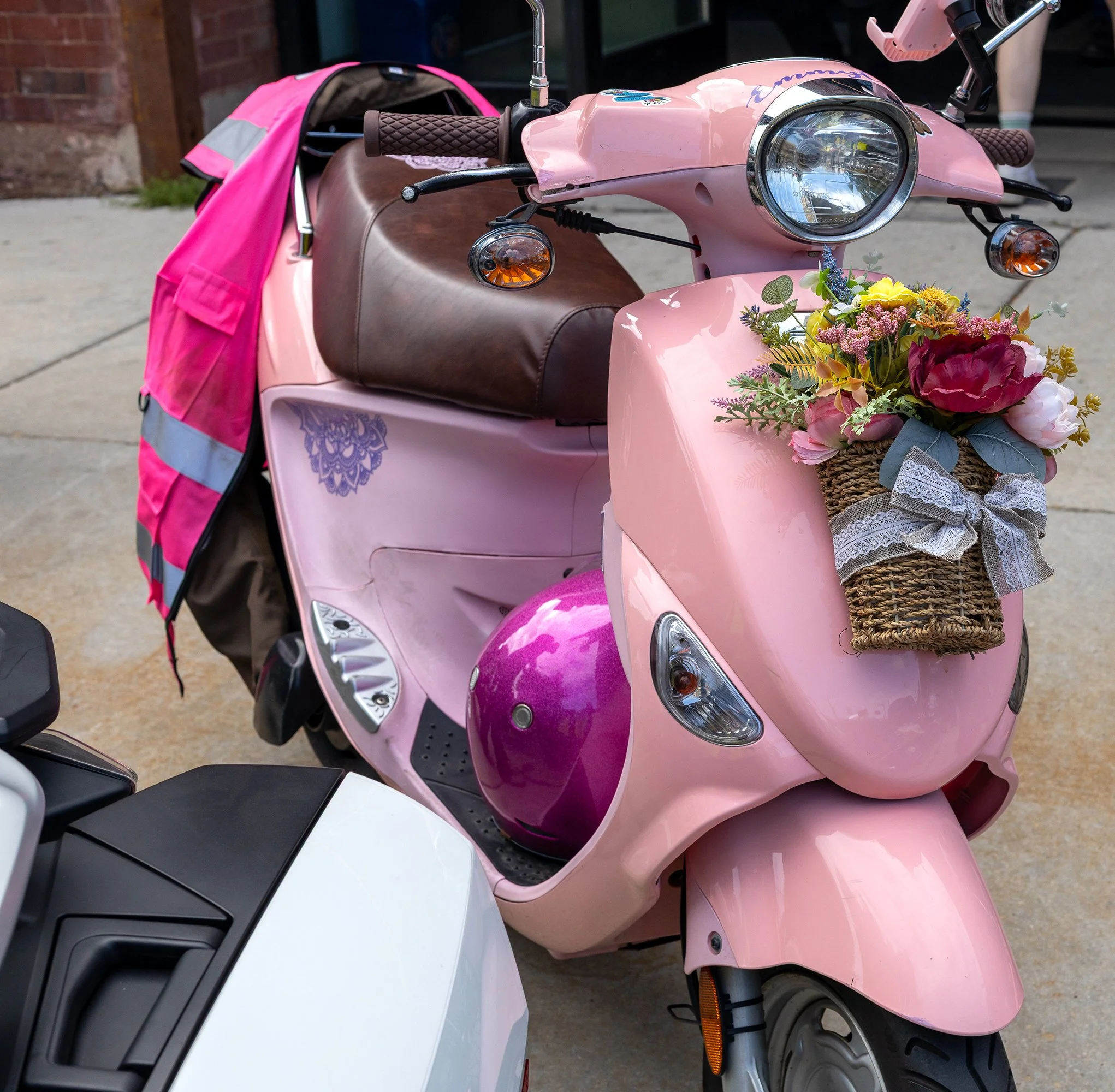 Close-up of two parked scooters, one pink with flowers in a basket at the front, the other white, on a sidewalk.