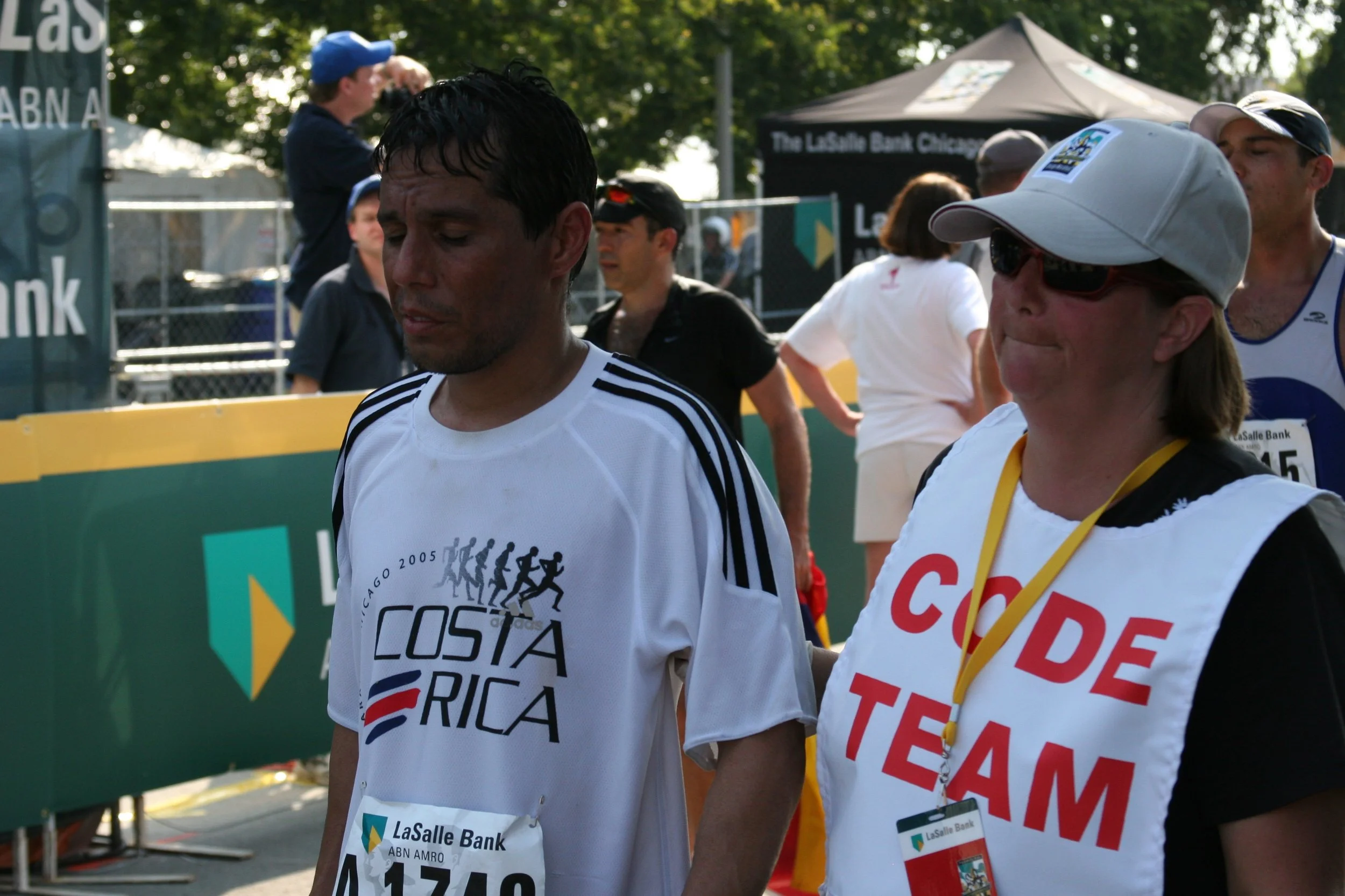 Two marathon runners standing side by side after race, wearing medals and running bibs with sponsor logos, with others and event tents in the background.