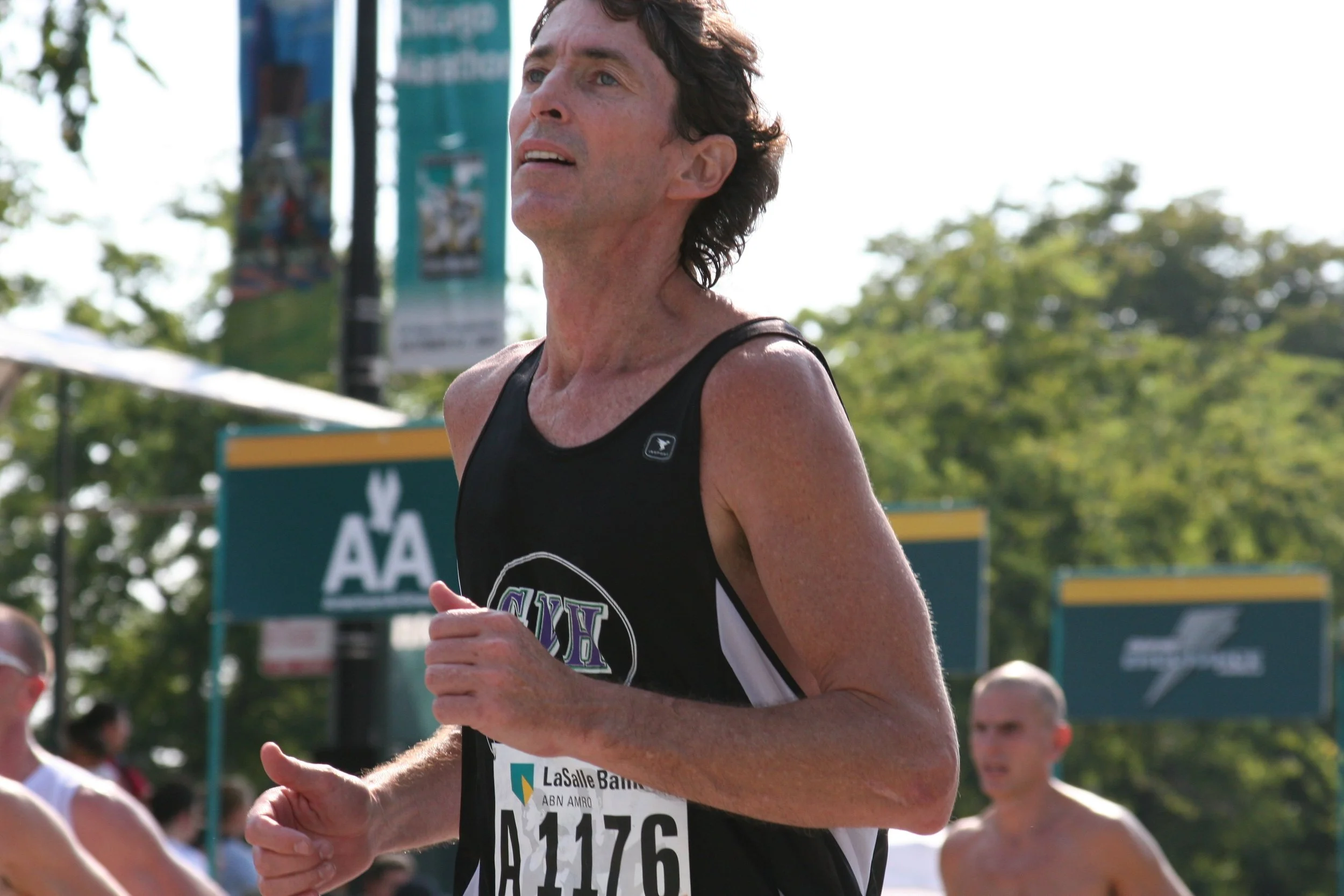 A male runner with a race bib number 11176, participating in a race outdoors on a sunny day. He is wearing a black athletic tank top with the words 'SIX' on it and has short dark hair. Other runners and trees can be seen in the background.