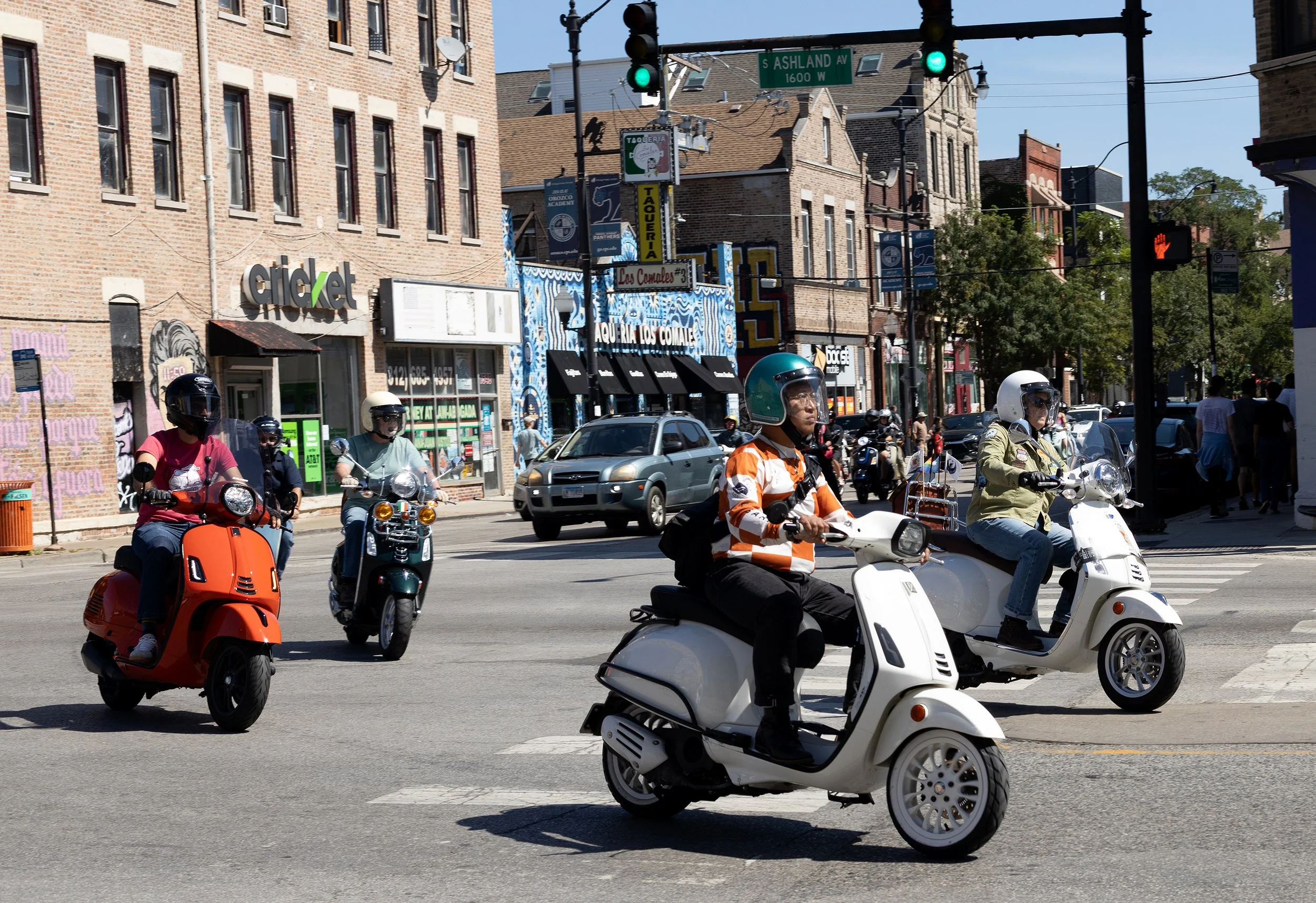 A busy urban intersection with multiple people riding scooters and motorcycles, a few cars, and various buildings in the background including storefronts and a street sign reading 'S Ashland Ave'.