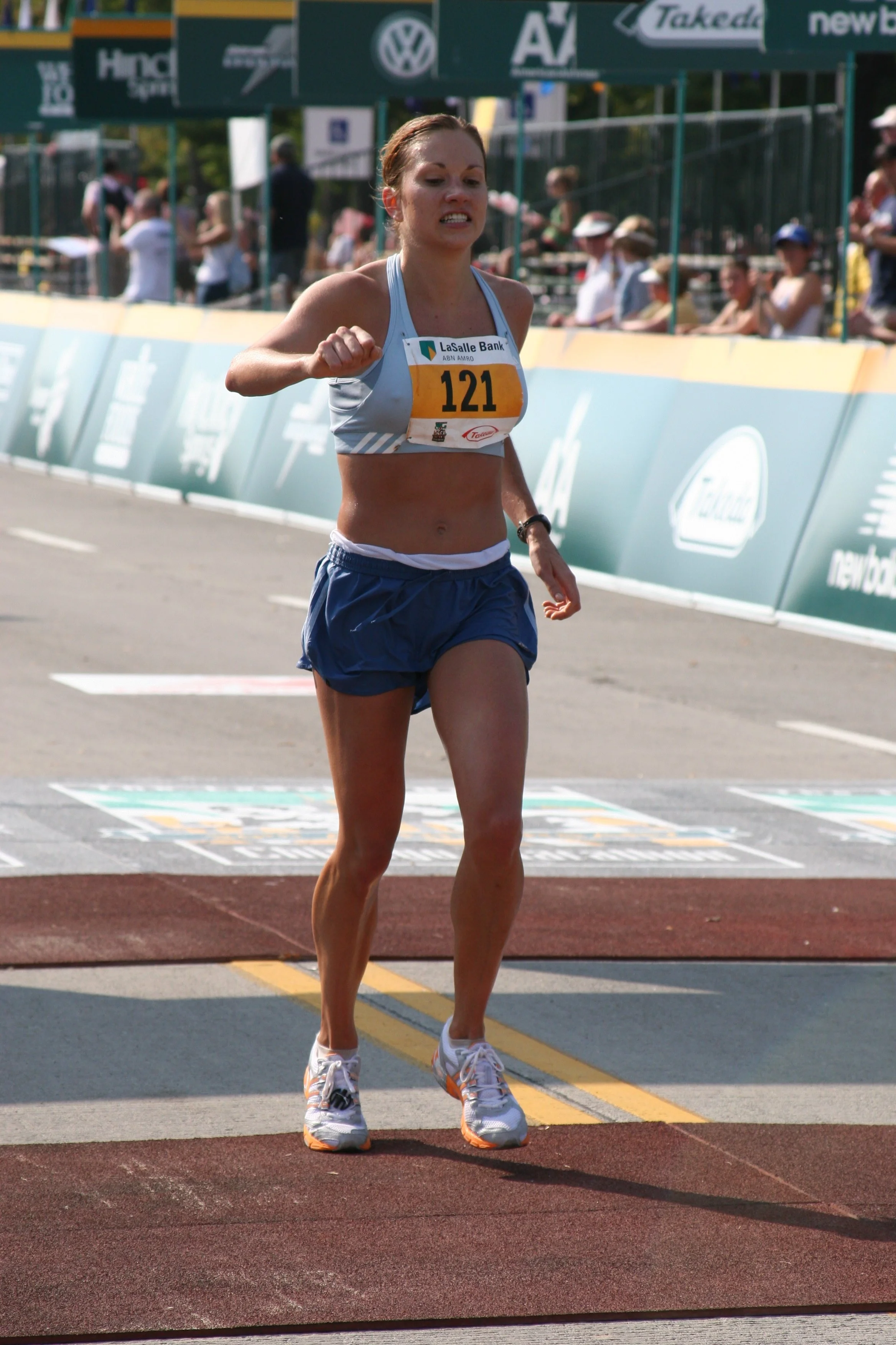 A female marathon runner in athletic gear, wearing a race bib with the number 121, finishing a race on a track with a crowd and banners in the background.