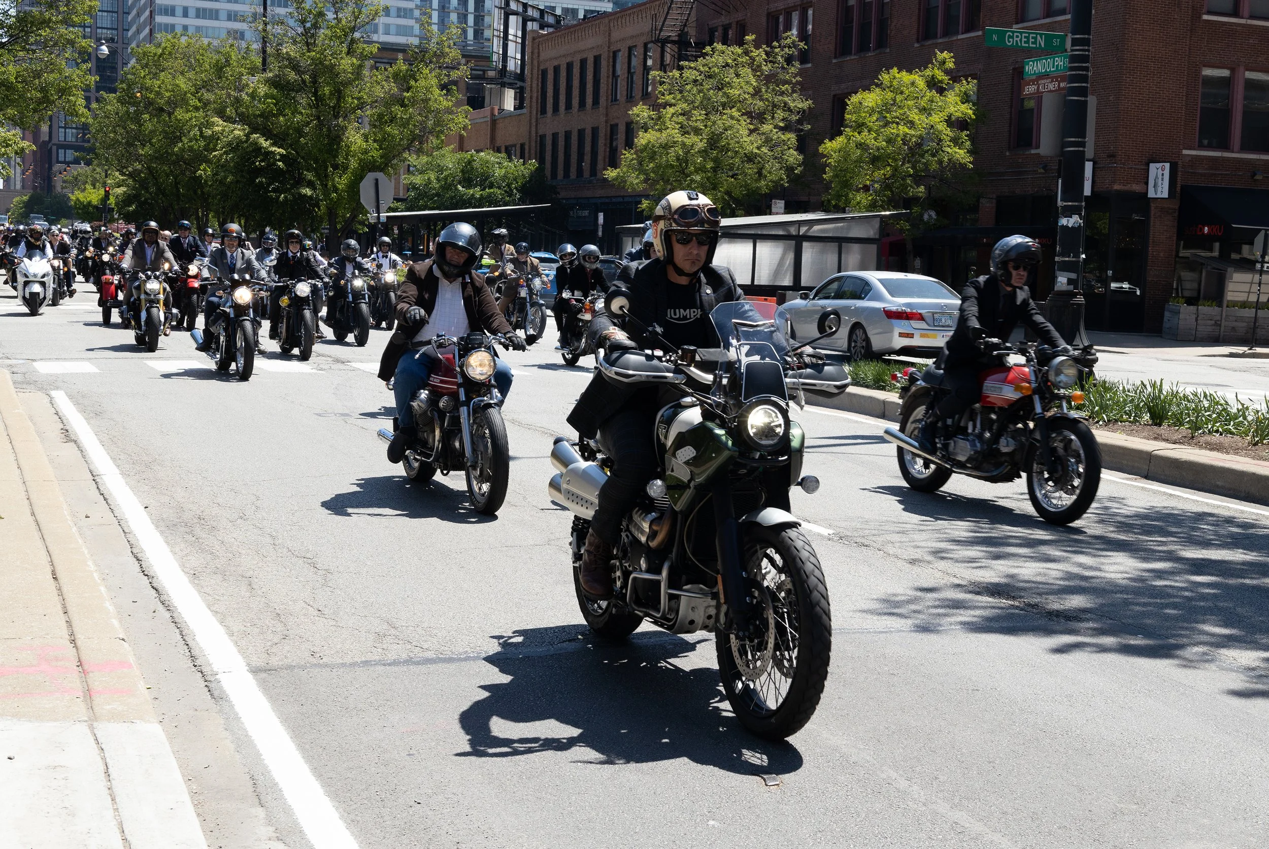 A large group of motorcyclists riding on an urban street, with some wearing helmets and sunglasses, surrounded by city buildings and cars parked along the side.