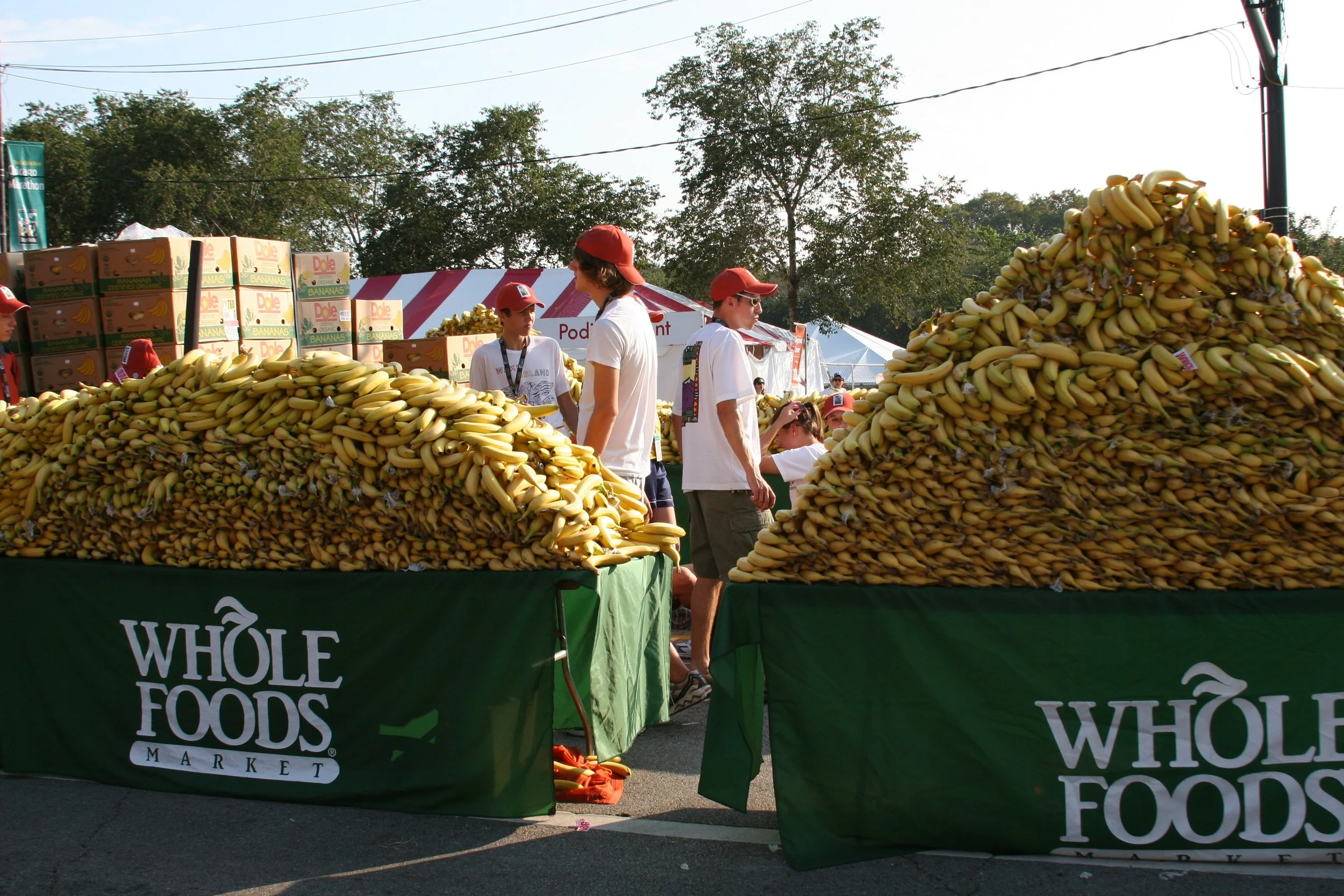 Market stalls stacked with bananas at an outdoor Whole Foods Market event, with people browsing and trees in the background.