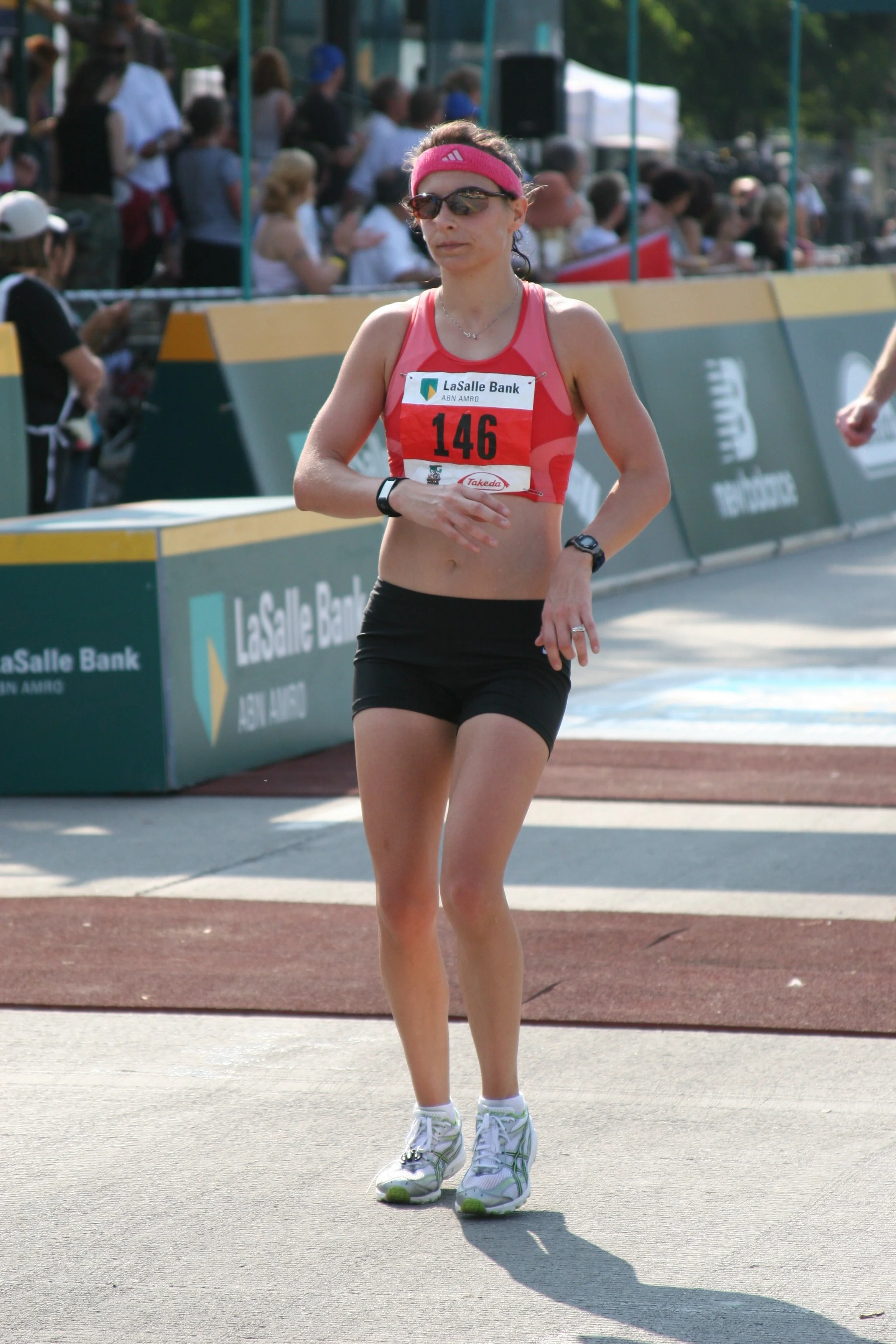 A woman running in a race wearing a pink headband, sunglasses, a red sports top with bib number 146, black shorts, and white sneakers, on a track surrounded by spectators and event banners.