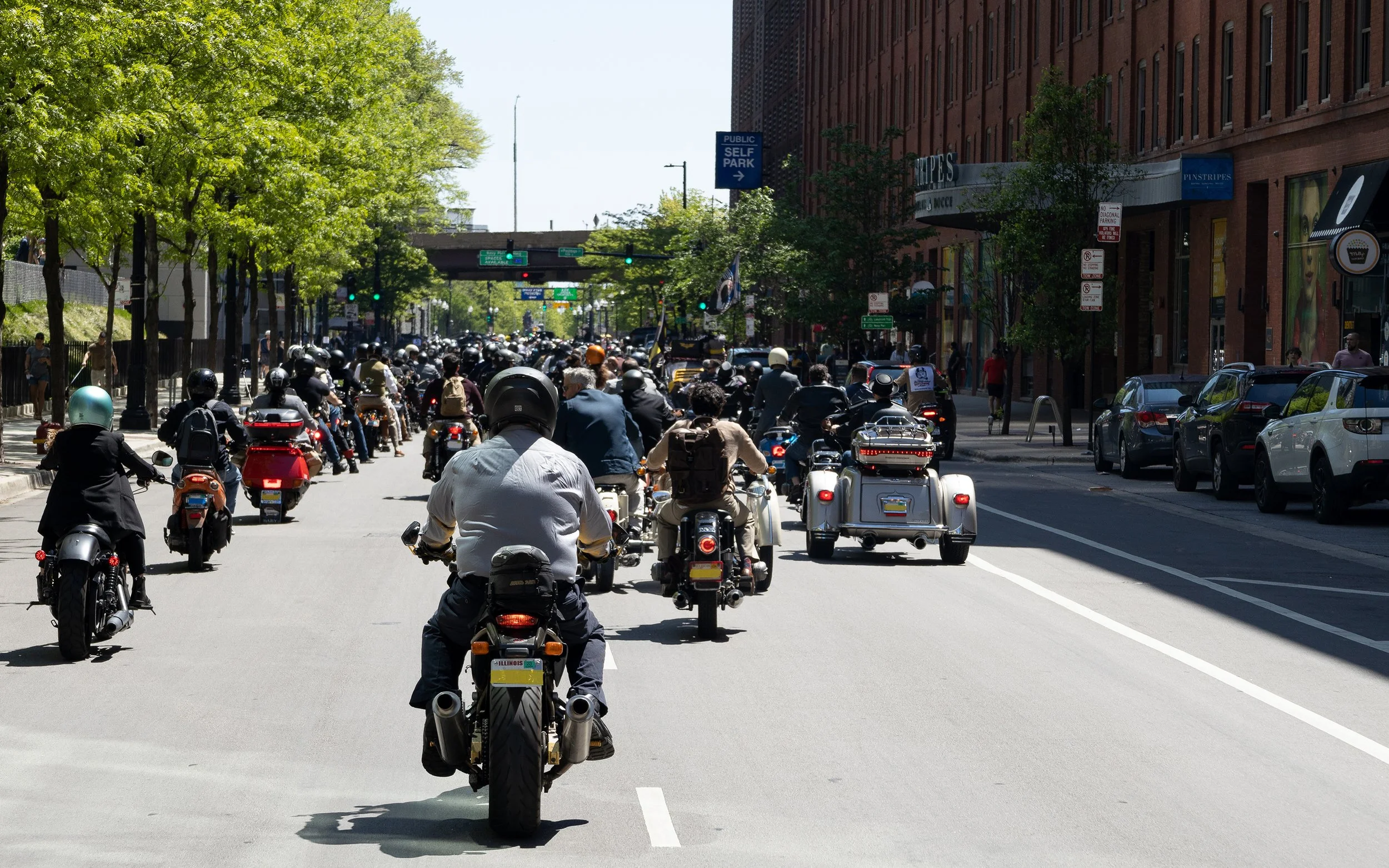 Traffic filled with motorcycles, scooters, and cars on an urban city street lined with trees and brick buildings.