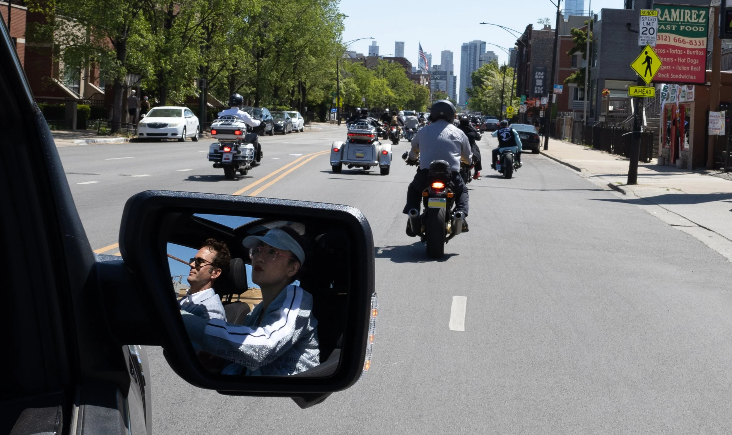 View from a vehicle's side mirror showing a woman and a man inside the vehicle. Beyond, a city street scene with motorcycles, cars, and pedestrians. Street signs and storefronts are visible with tall buildings in the background.