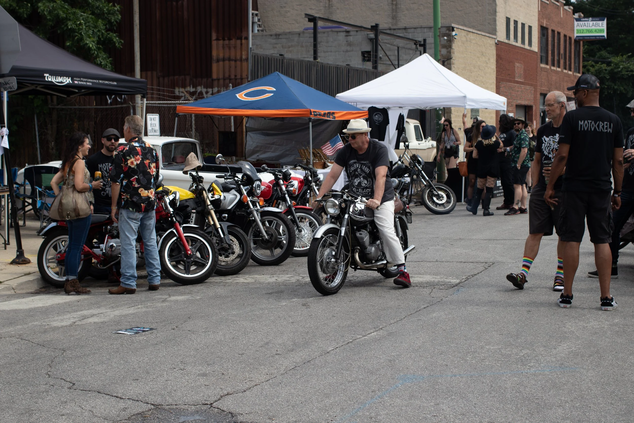 People at an outdoor event with motorcycles and tents, some talking and others walking, in a street scene with buildings in the background.
